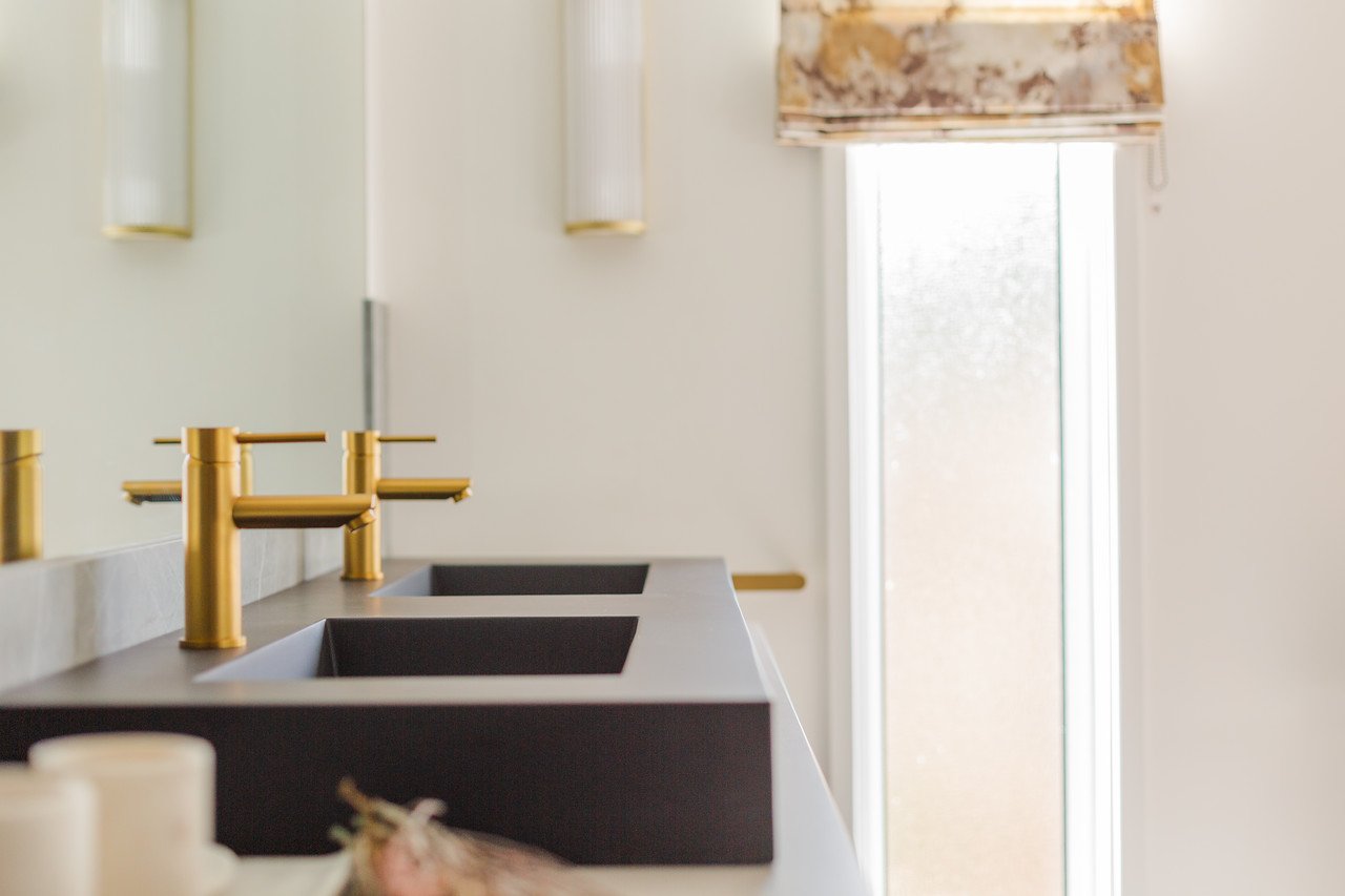“Modern wall light in the Gamlingay ensuite bathroom, illuminating a double‑sink vanity with black basins and gold fixtures