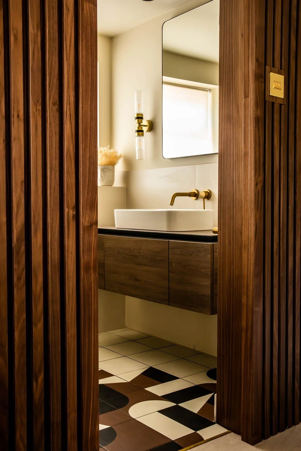 Contemporary bathroom with a floating wooden vanity, white vessel sink, brass wall‑mounted tap and warm timber slatted doorway.