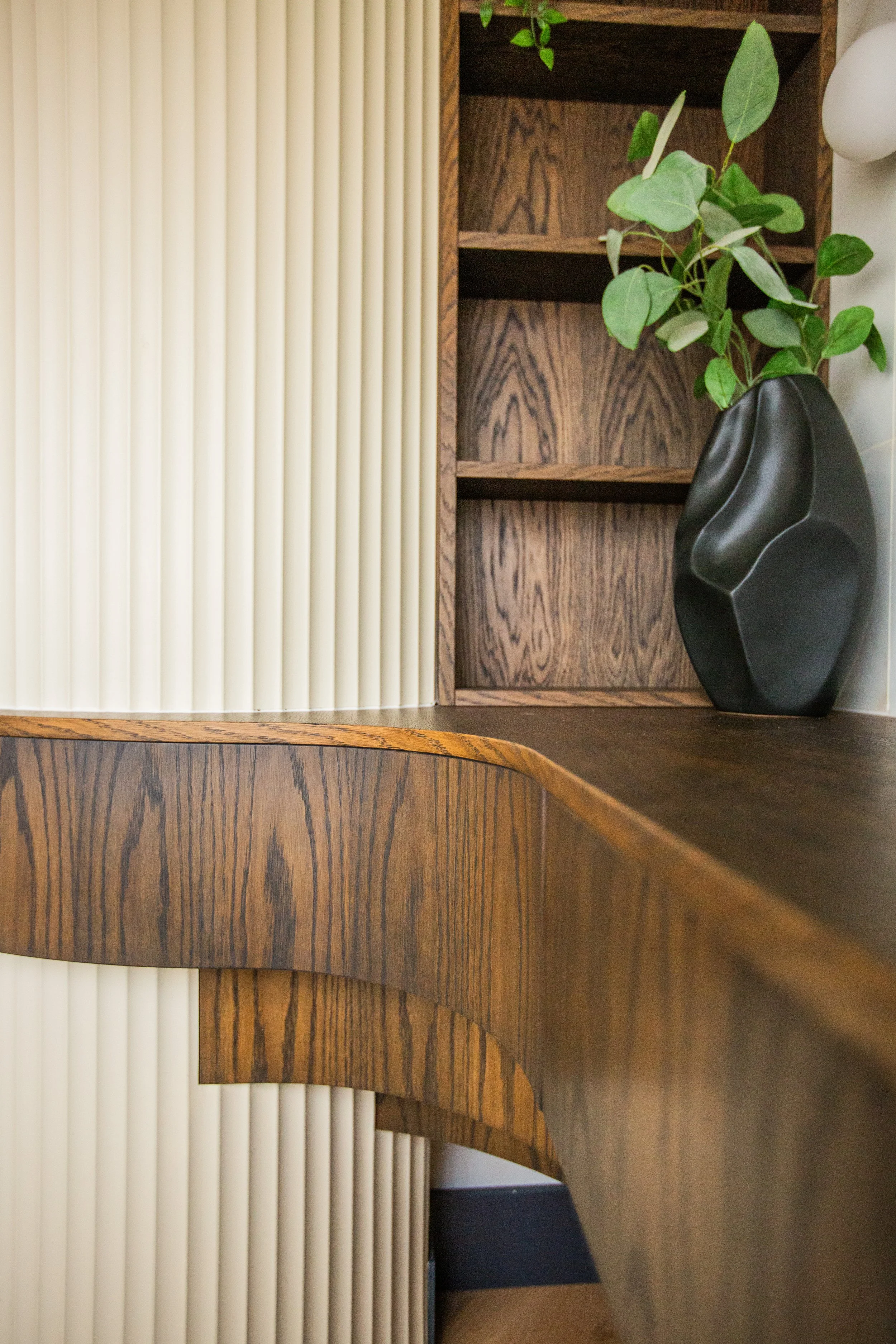 Curved wooden countertop with fluted wall panel, dark timber shelving, and a sculptural vase with greenery in a contemporary interior