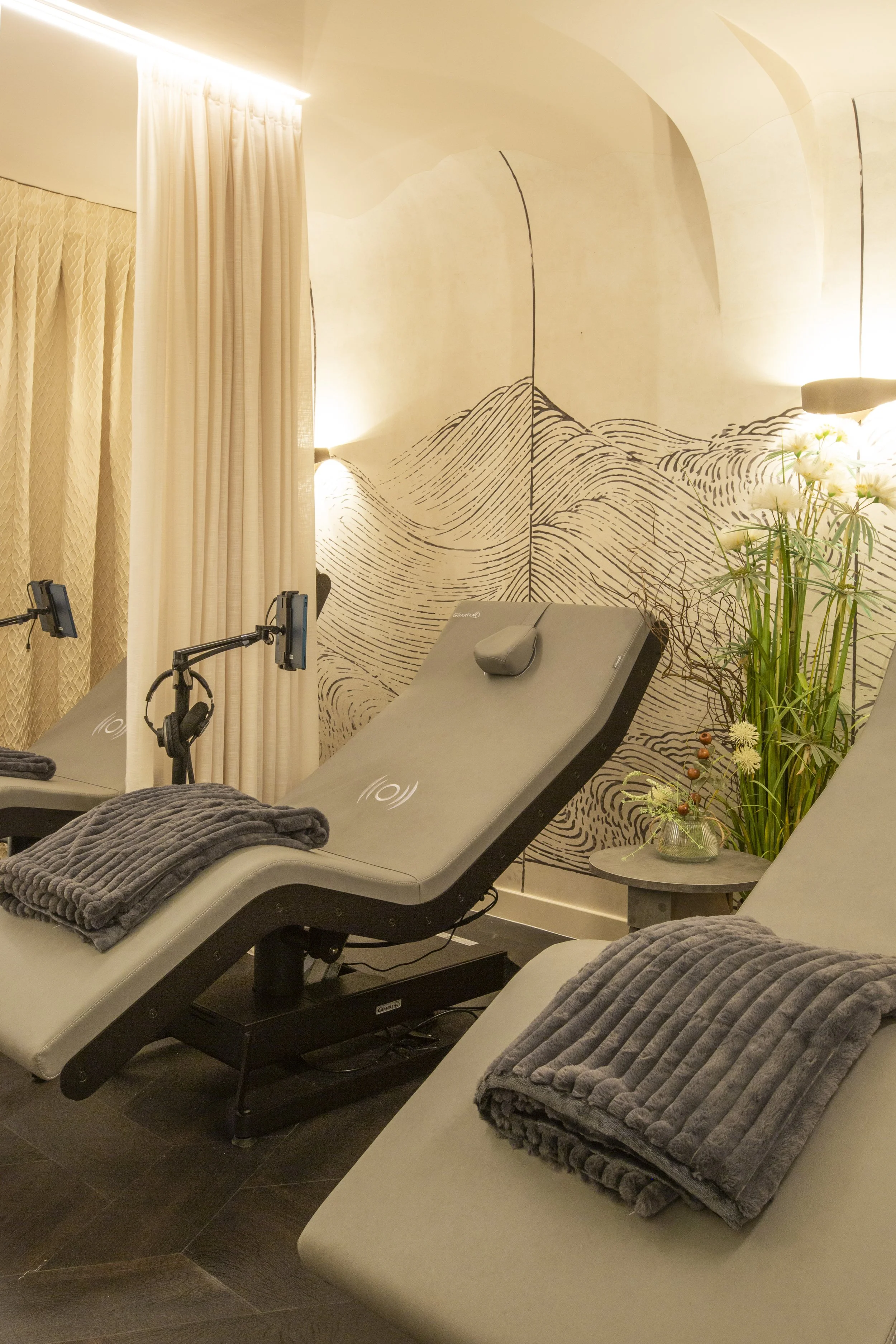 “Spa treatment room with two reclining lounge chairs, soft grey blankets, a stylised mountain mural, warm lighting, and natural greenery.”