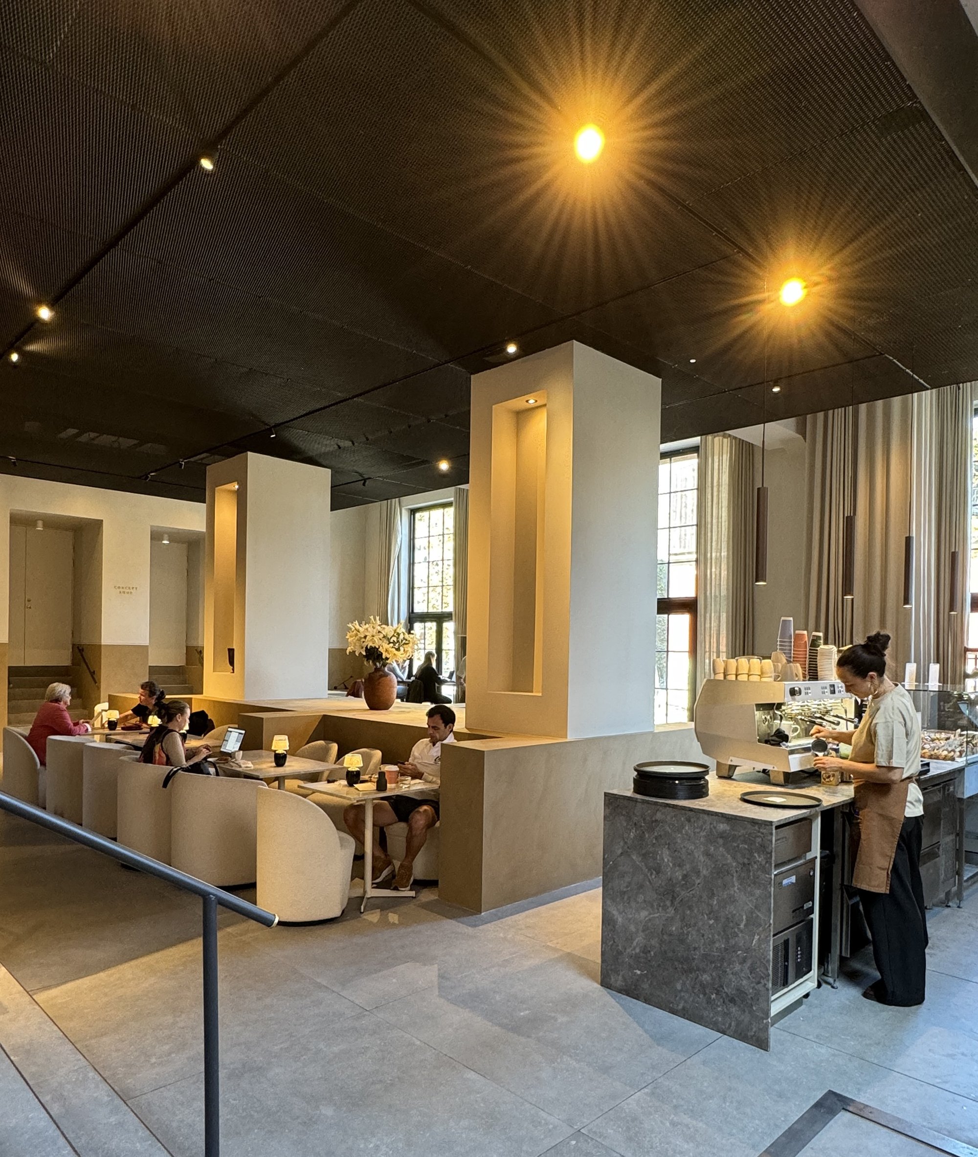 Moderne caféinteriør med gæster siddende ved borde, en barista ved kaffemaskine, dæmpet belysning og store vinduer.