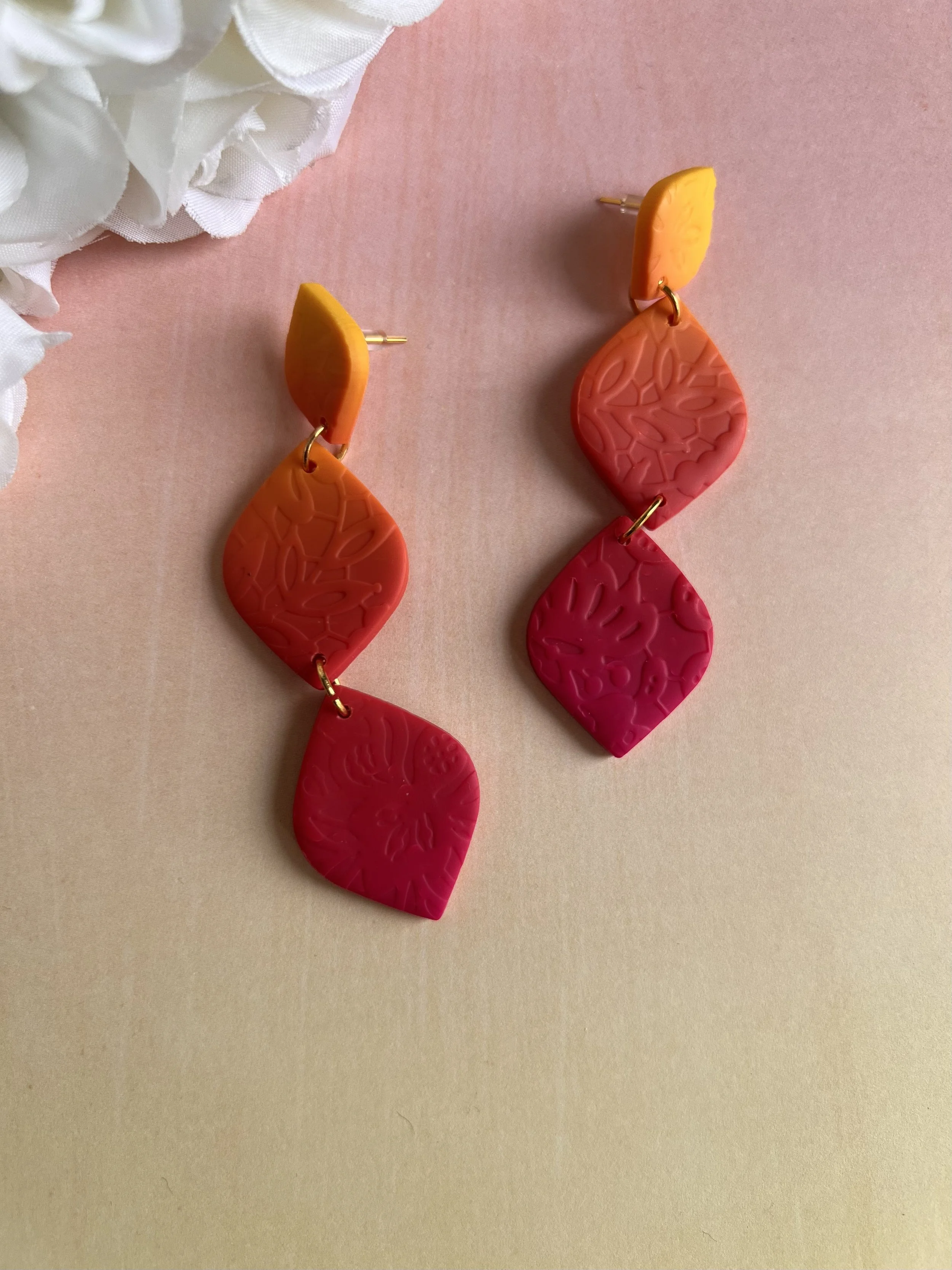 Pair of colorful, abstract earrings with pink, red, orange, and yellow palette, placed on a light pink surface near a white flower in the corner.