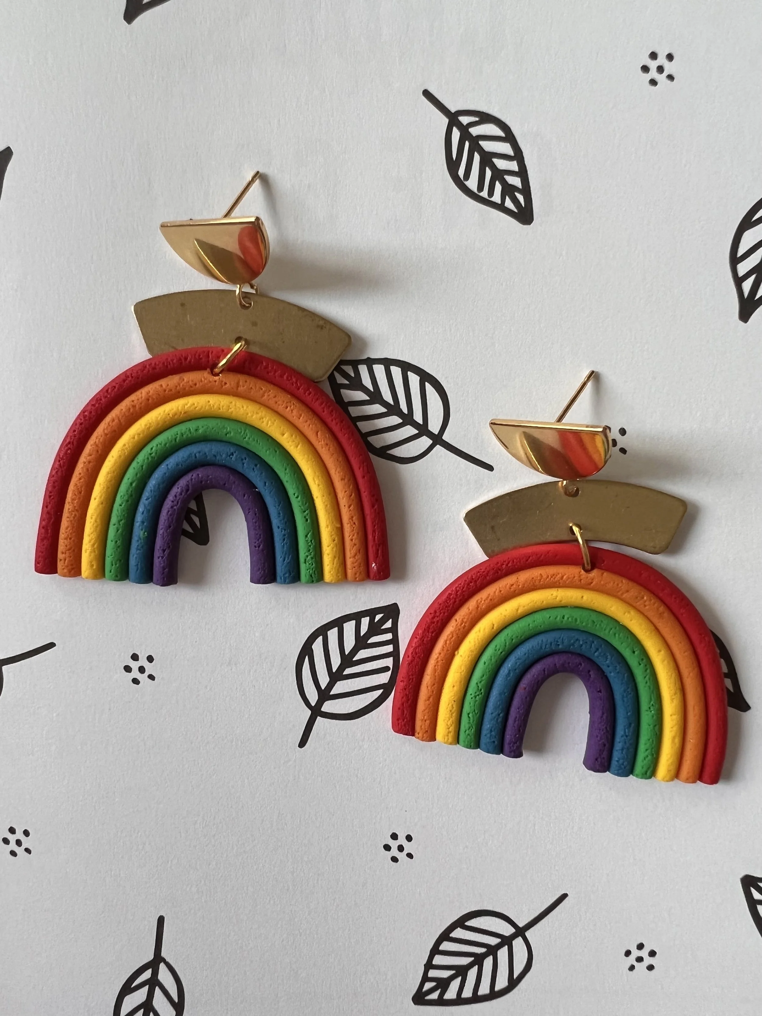 Colorful rainbow-shaped earrings with gold half-circle studs on a white background with black leaf illustrations.