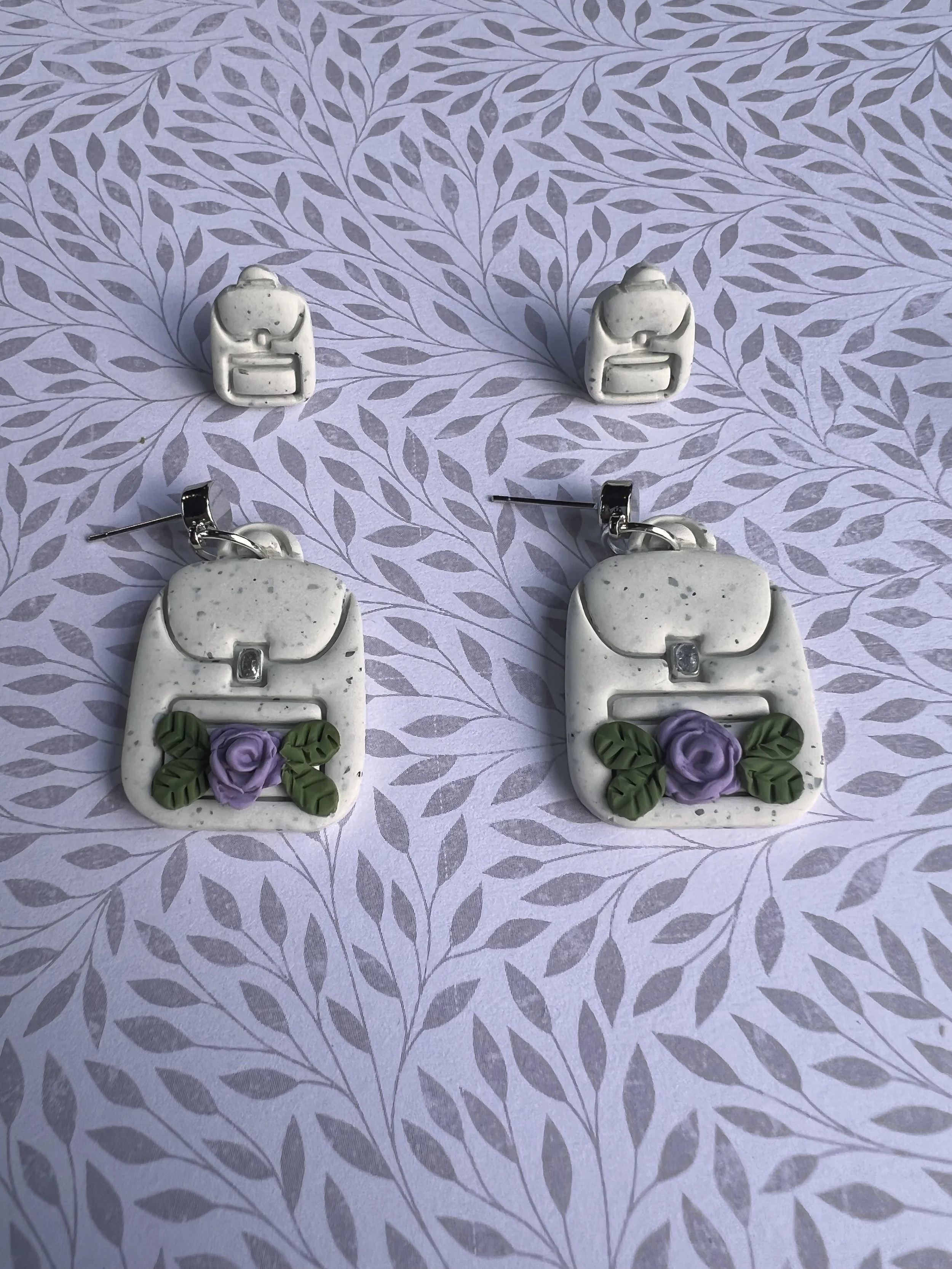 Pair of earrings and matching stud earrings shaped like handbags with purple roses and green leaves on a patterned purple background.