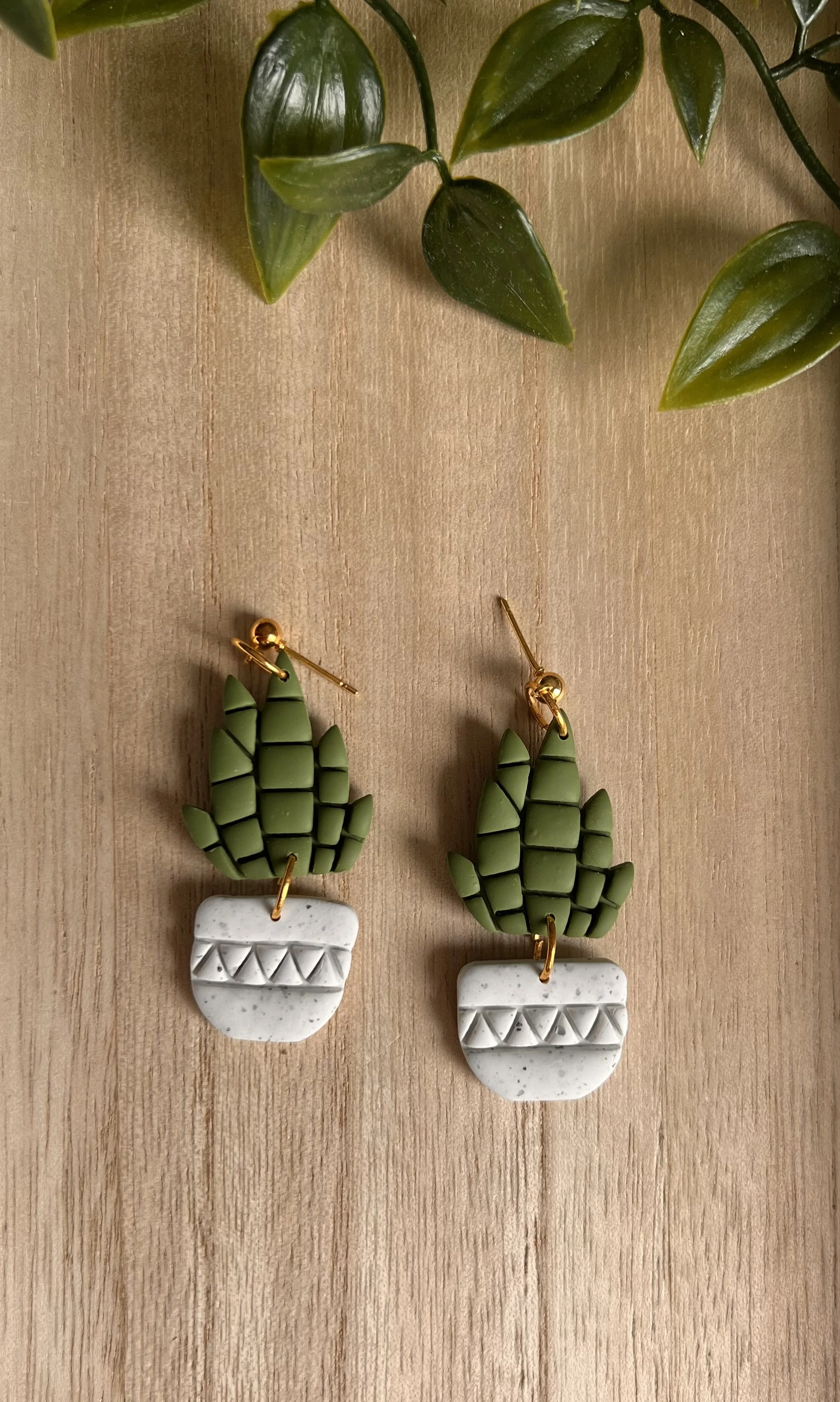 Pair of cactus-shaped earrings with green and white geometric design, hanging on a light wooden surface, with green plant leaves partially visible at the top.