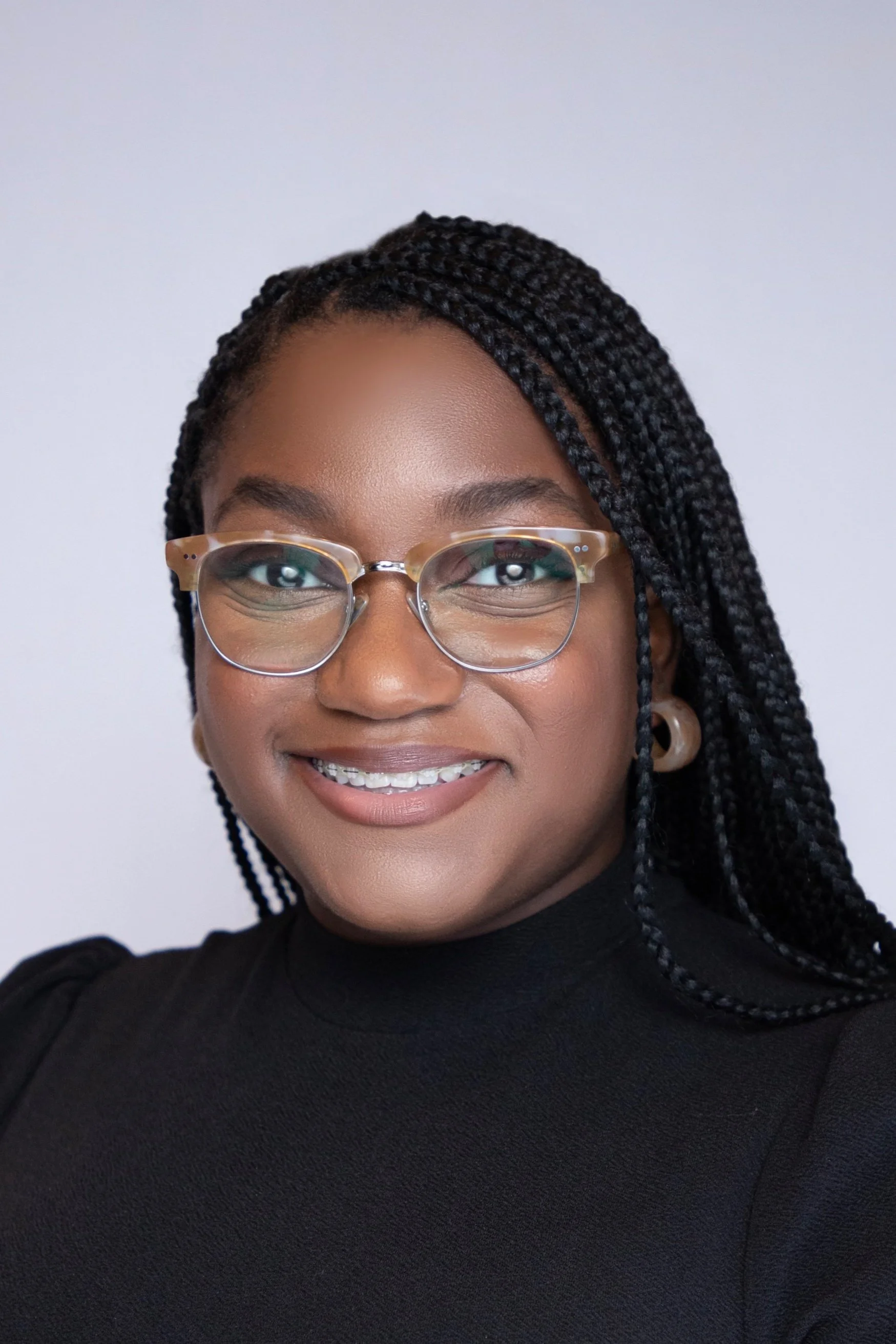 A smiling woman with black braided hair, glasses, hoop earrings, and a black top against a plain light background.