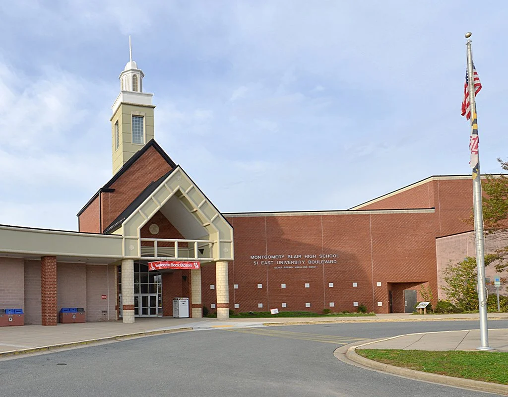 The exterior of Montgomery Blair High School, a brick building with a tower, situated on East University Boulevard with a flagpole and American flag.
