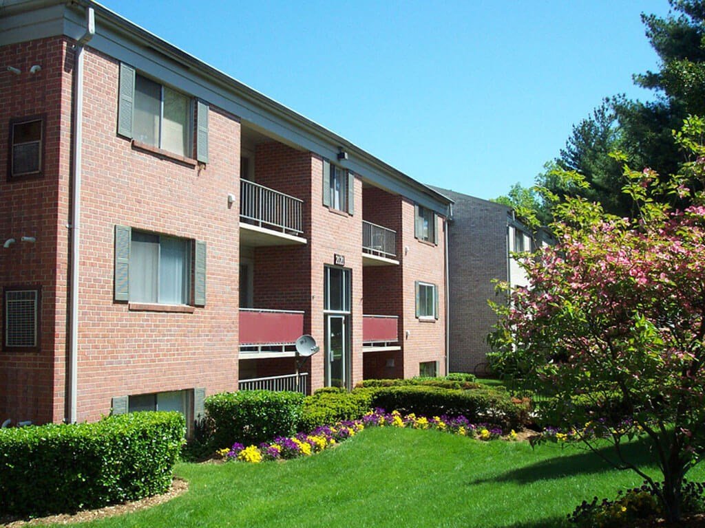 A brick apartment building with three floors, balconies, and lush landscaping including trimmed bushes, colorful flowers, and a flowering tree under a clear blue sky.