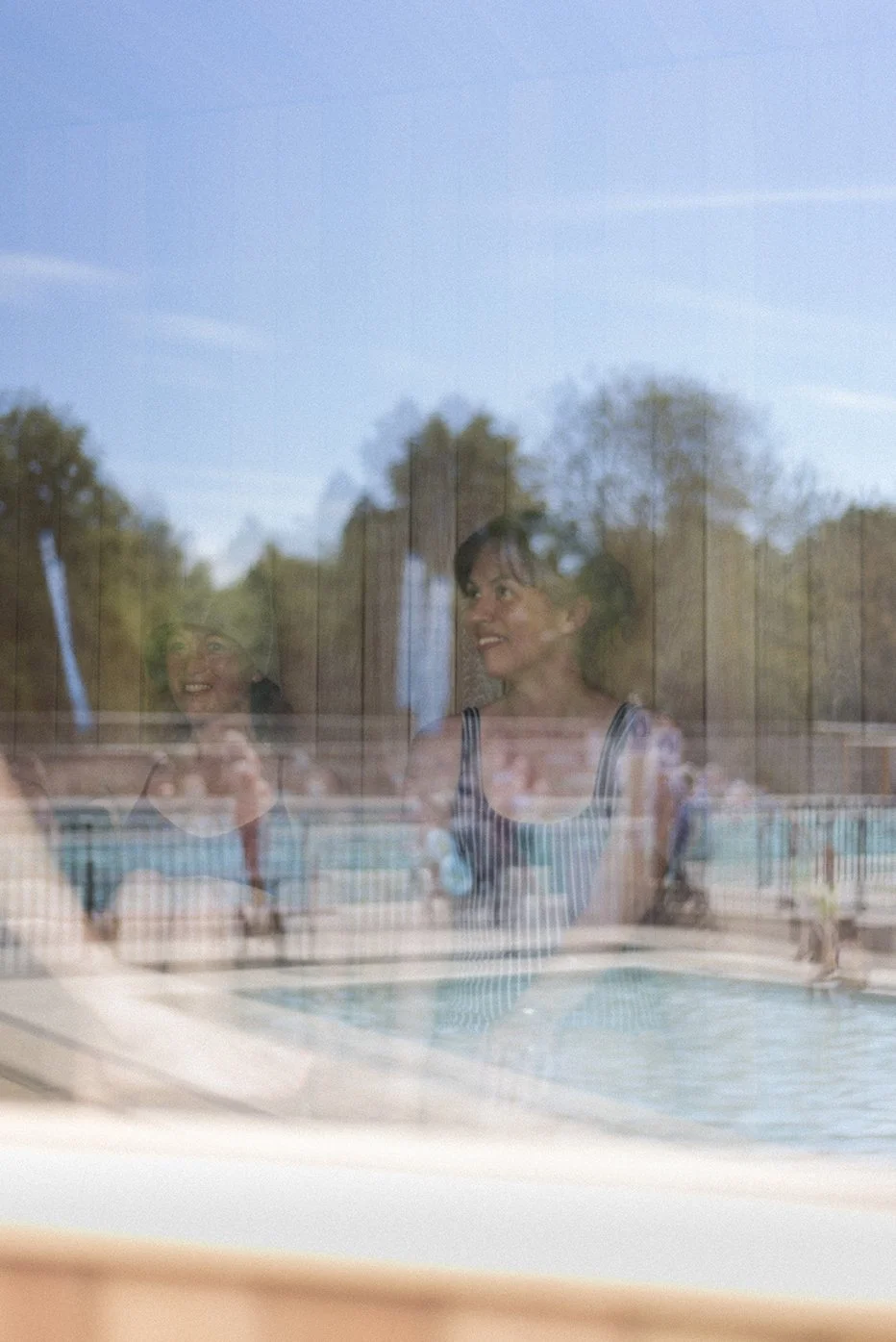 Two women smiling and talking, seen through a sauna window, with a swimming pool and trees in the background.