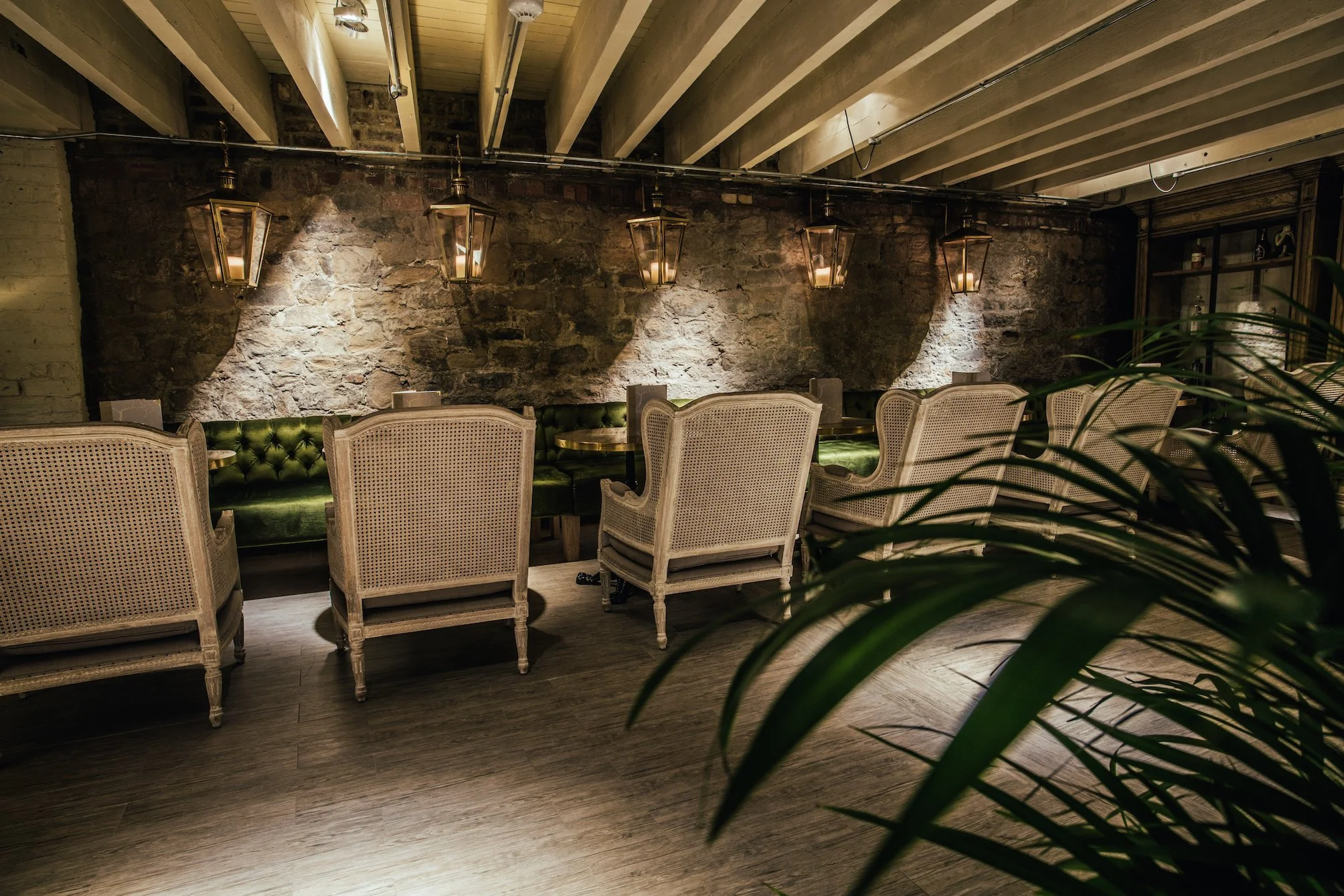Interior of a cozy restaurant with a stone accent wall illuminated by warm pendant lights, vintage-style chairs with wicker backs and green velvet seats arranged around tables, and a dark wooden floor, with greenery partially visible in the foreground.