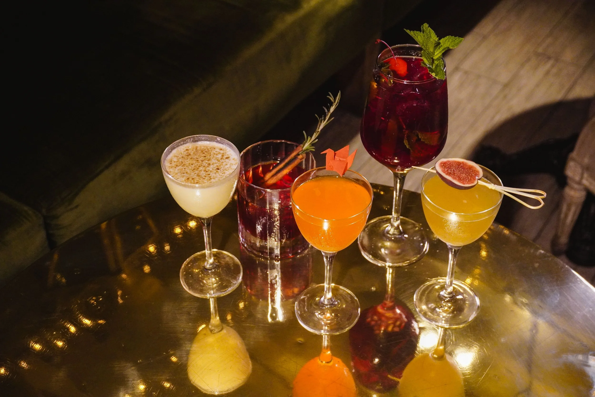 Assorted cocktails on a reflective golden table, garnished with fruit, herbs, and decorative elements, set against a dark background.
