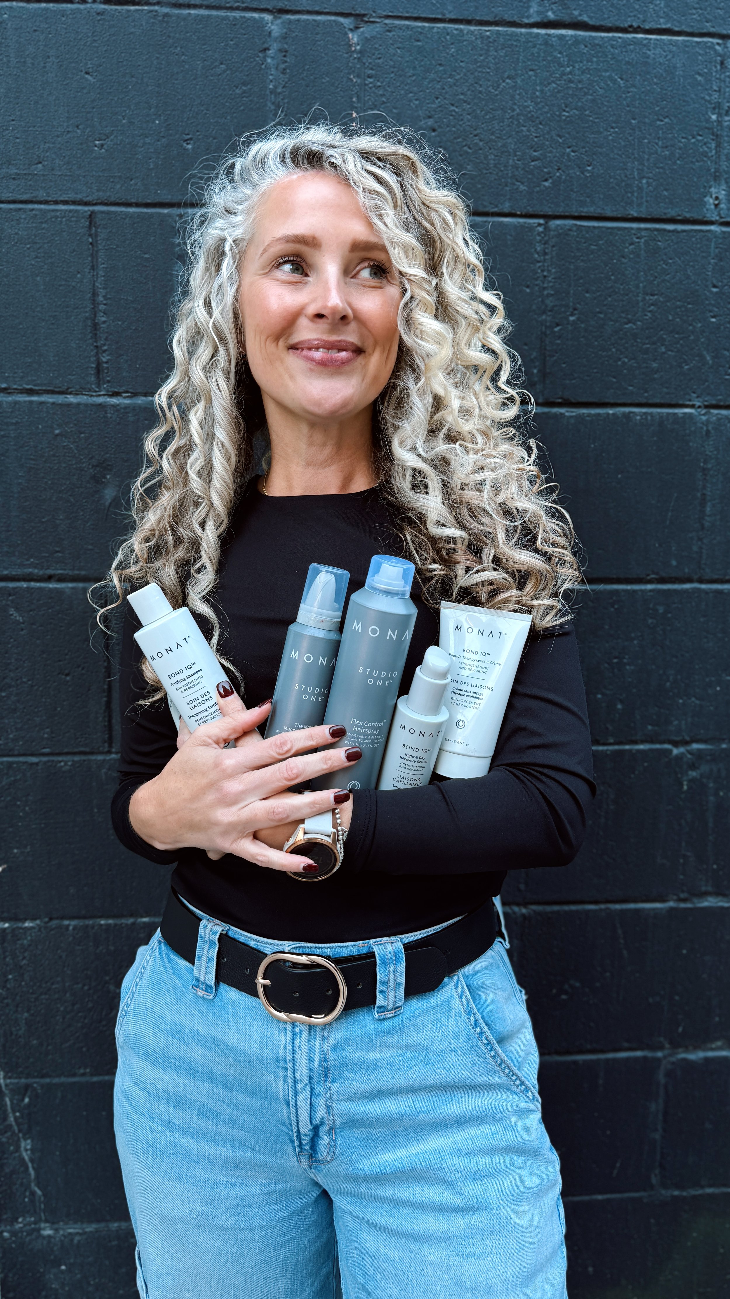 A woman with curly blonde hair holding several hair care products in front of a black brick wall, wearing a black long sleeve shirt, blue jeans, a black belt with a silver buckle, and a wristwatch.