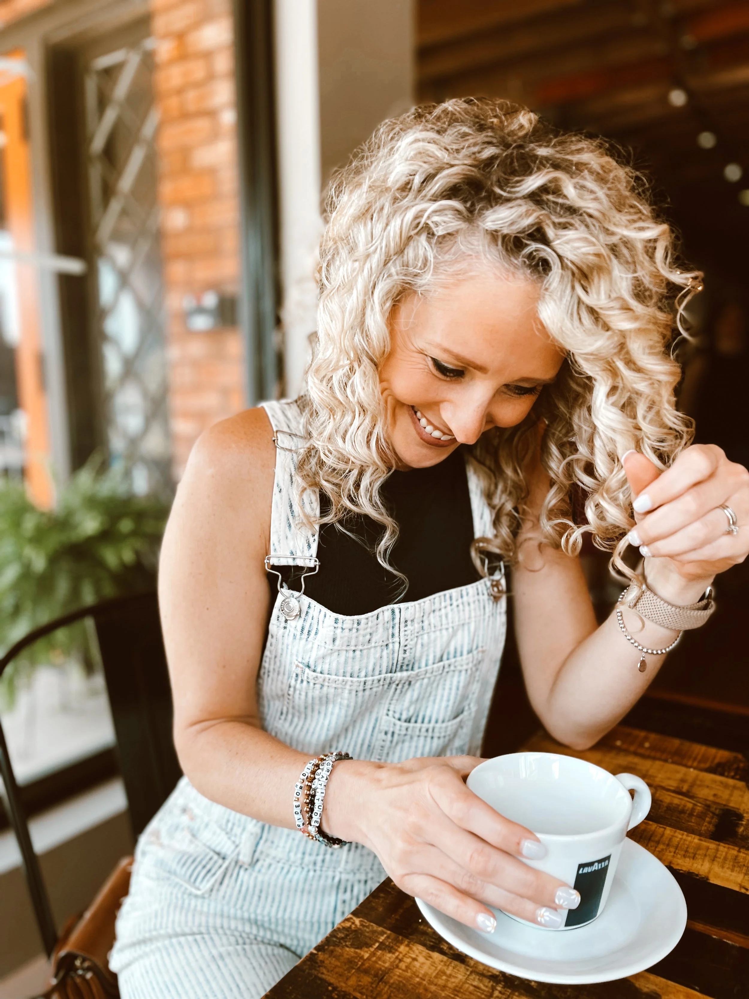 A woman with curly blonde hair sitting at a wooden table, holding a white coffee cup in a saucer, smiling and looking down at the cup.