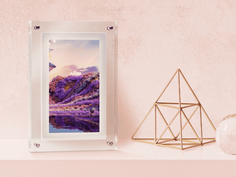 A digital photo frame displaying an artistic landscape with purple foliage on a pink backdrop, next to a gold geometric sculpture on a light pink surface.