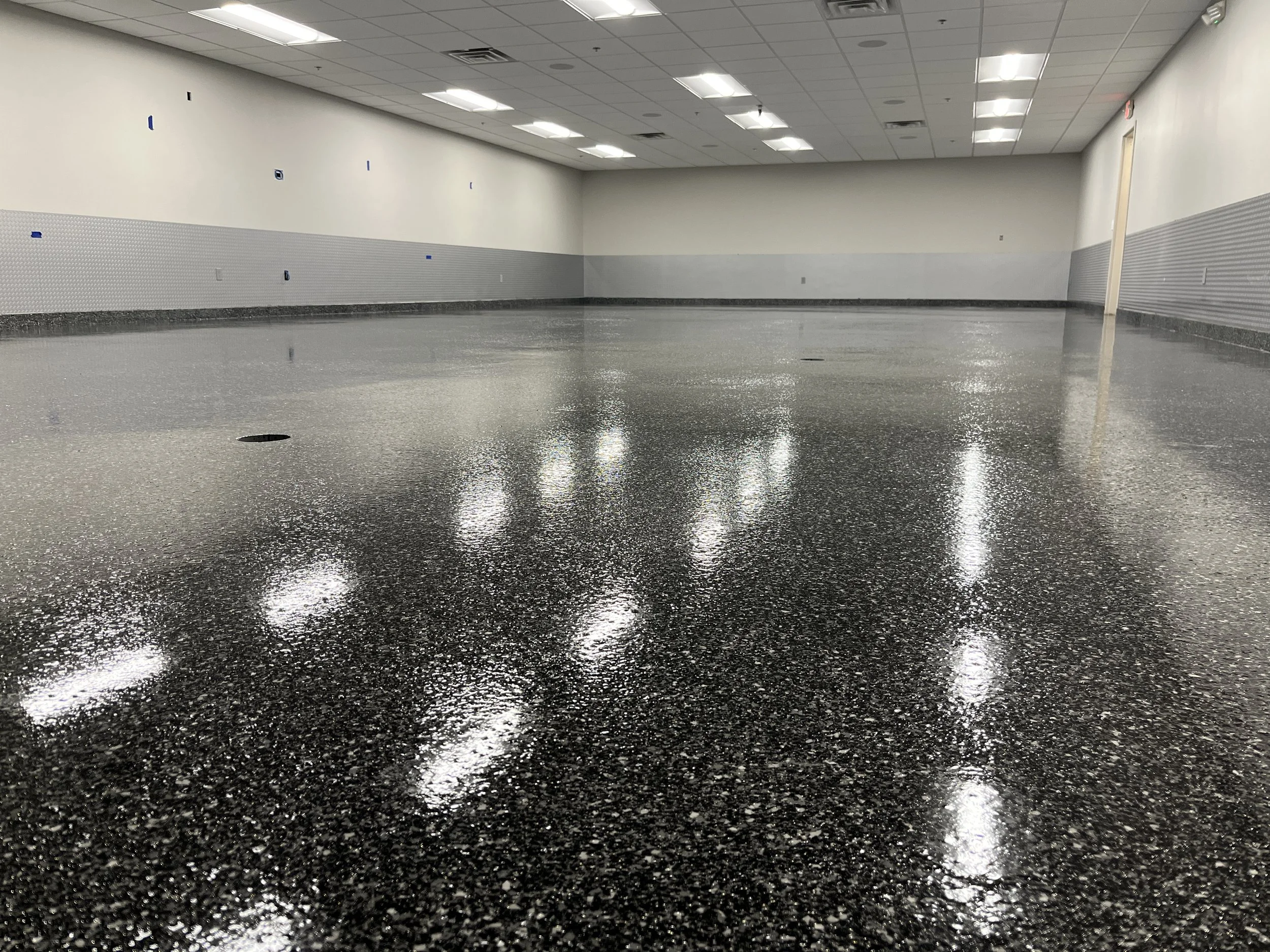 Newly finished black speckled epoxy floor in a large, well-lit room with white walls and ceiling, and fluorescent lights reflecting on the surface.