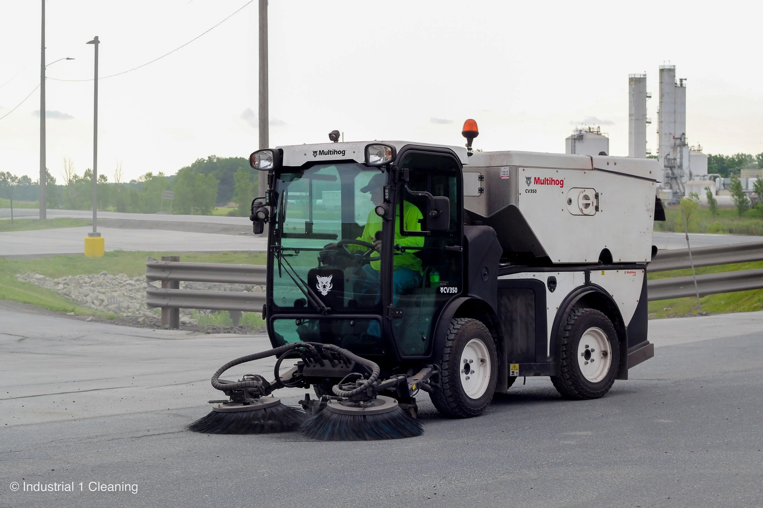 A street sweeper vehicle with dual rotating brushes cleaning a paved road, with industrial buildings and trees in the background.