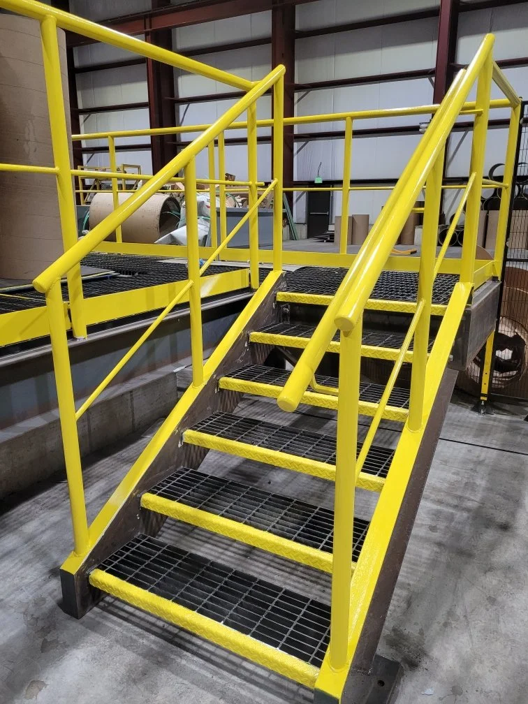 Yellow industrial safety staircase with handrails, metal steps, and grated treads leading to an elevated platform in a warehouse setting.