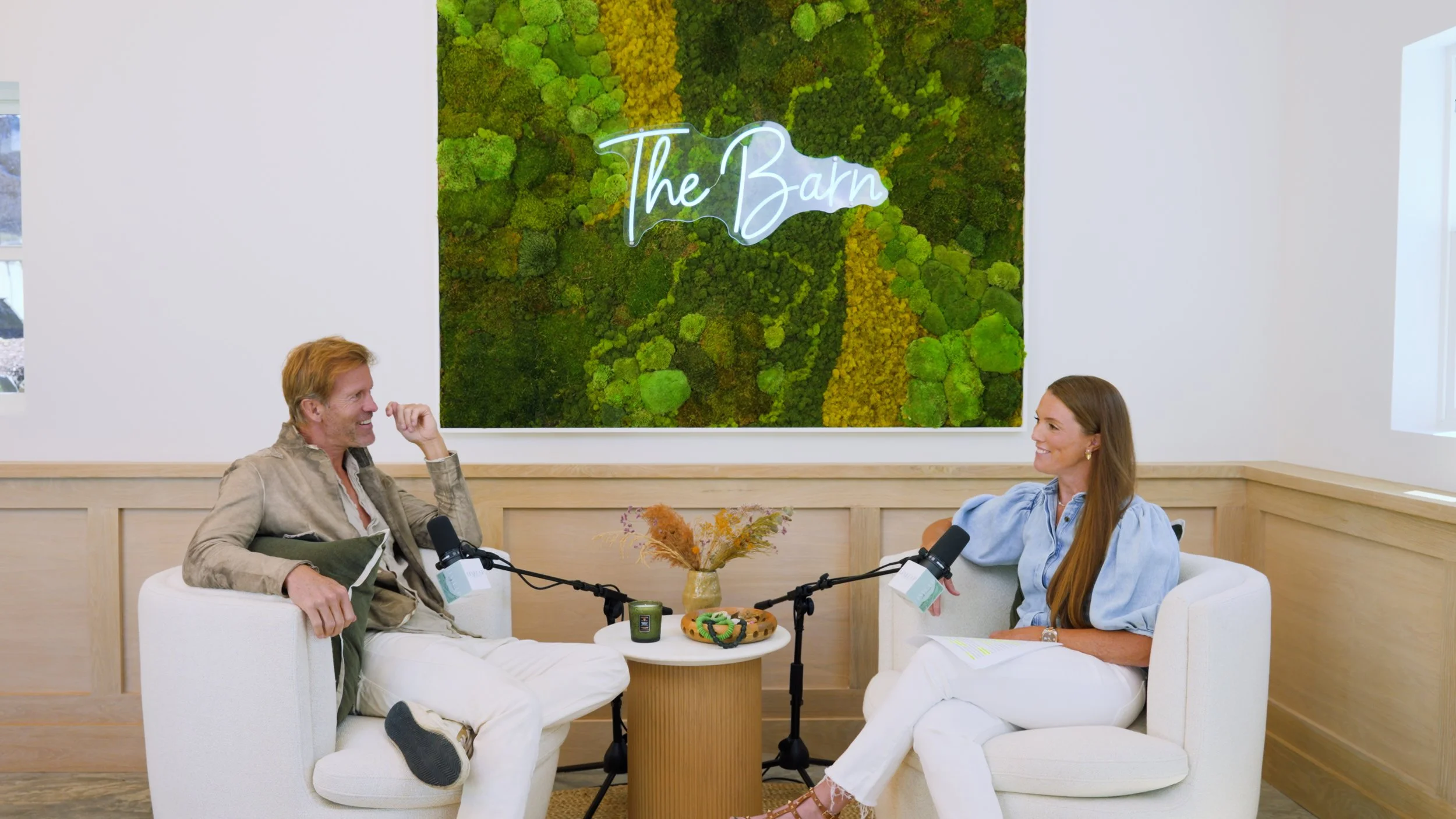 Two people, a man and a woman, are sitting in white armchairs having a conversation with microphones in front of them. There is a small round table between them with a vase of dried flowers, a candle, and a decorative piece. Behind them is a large green moss wall with a neon sign that says 'The Barn.' The room has white walls, wood paneling, and a window on the right.