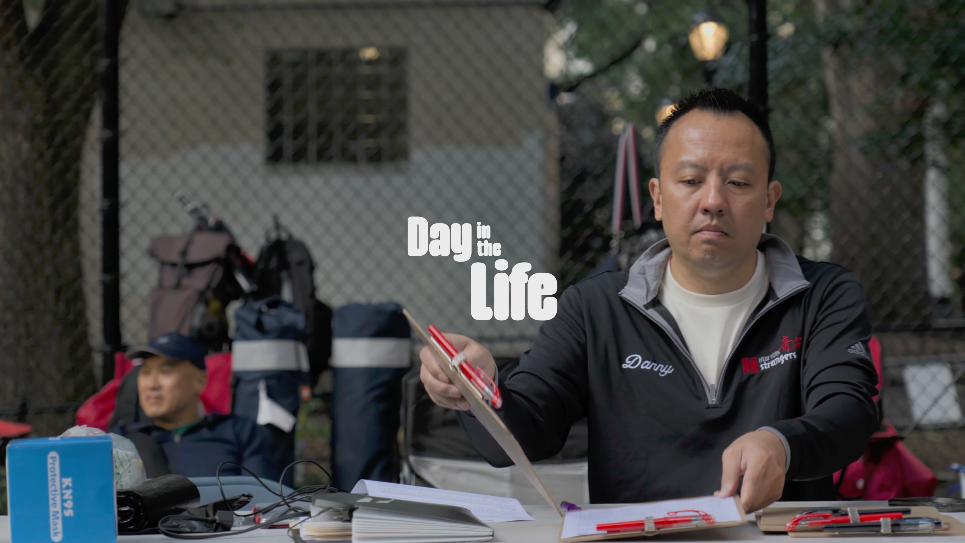 Man sitting at a table outdoors, reading a book, with backpacks and supplies in the background, and the text 'Day in the Life' overlaid.