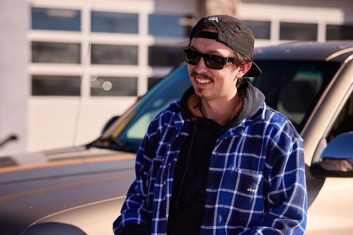 A young man with a mustache and beard wearing sunglasses, a backward baseball cap, and a blue checkered shirt, smiling while standing outdoors near a car with a building in the background.