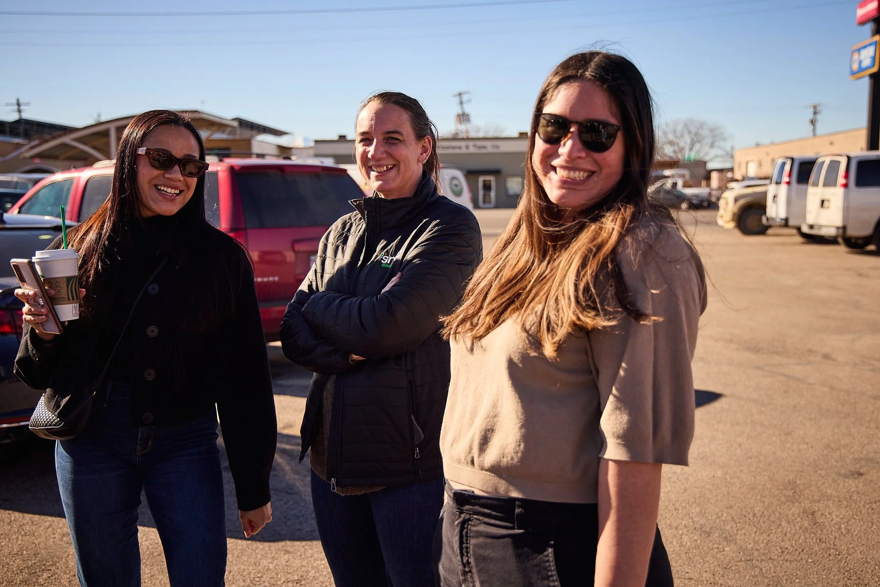 Three women standing together outdoors in a parking lot, smiling and looking at the camera, with cars and buildings in the background.