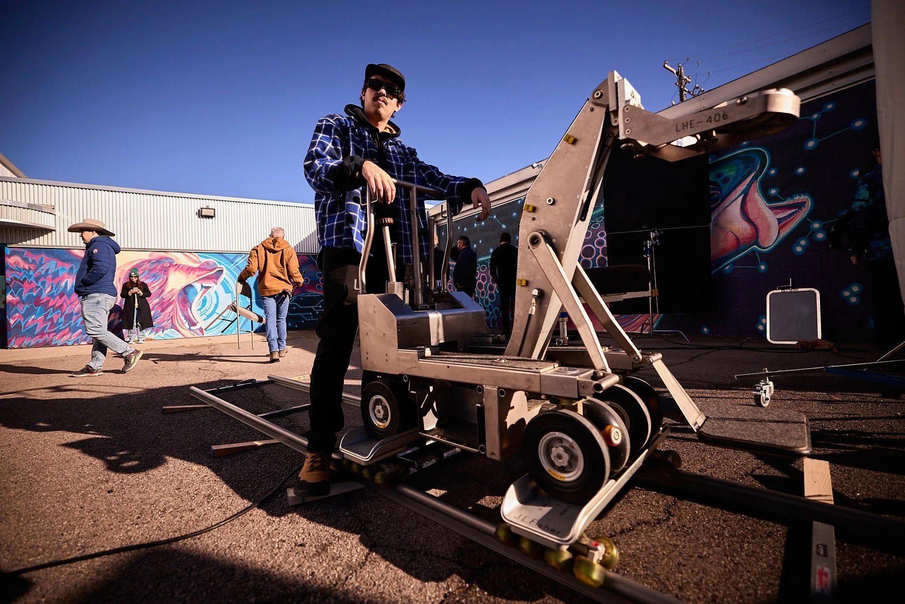 A person operating a film camera on a dolly on an outdoor set with graffiti art and a mural of a roaring lion on the wall behind them.