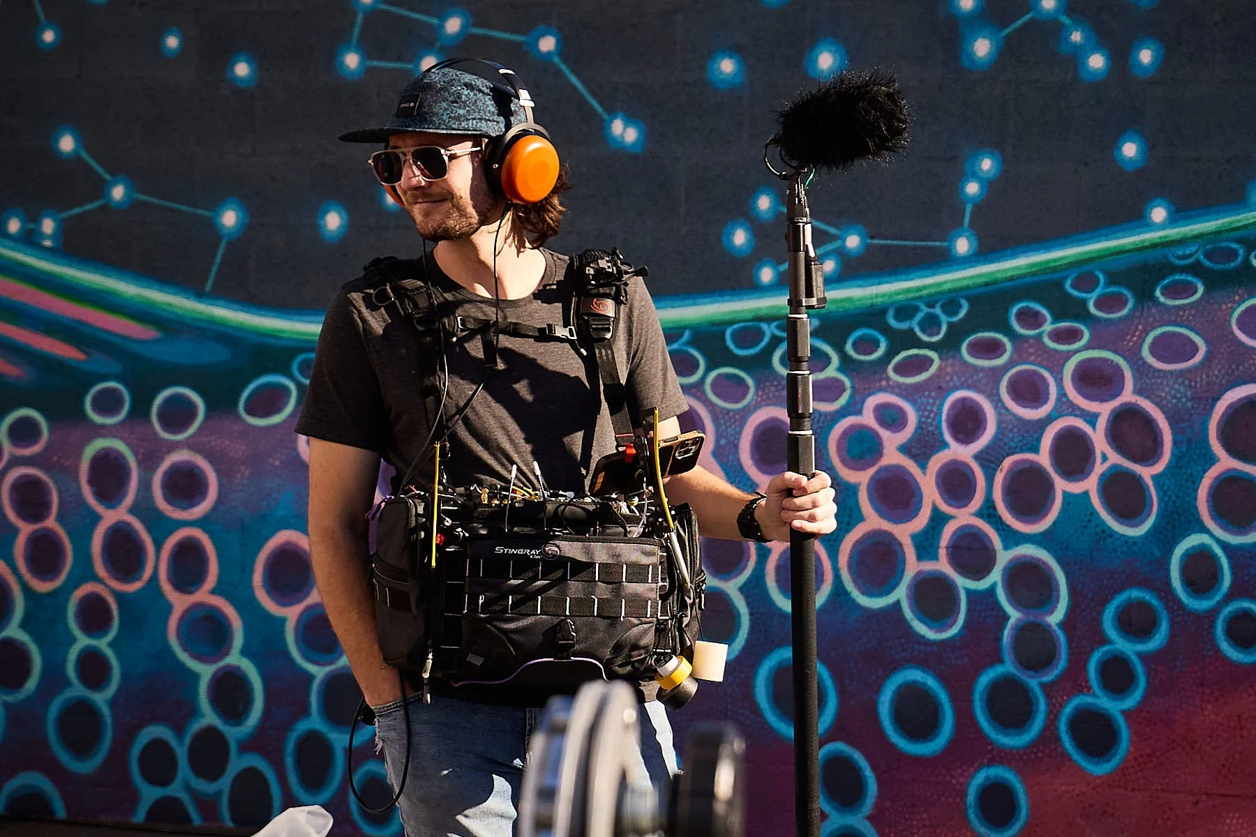 A man wearing sunglasses, a hat, and headphones stands outdoors against a colorful, abstract background, holding a boom microphone, and wearing audio equipment around his waist.