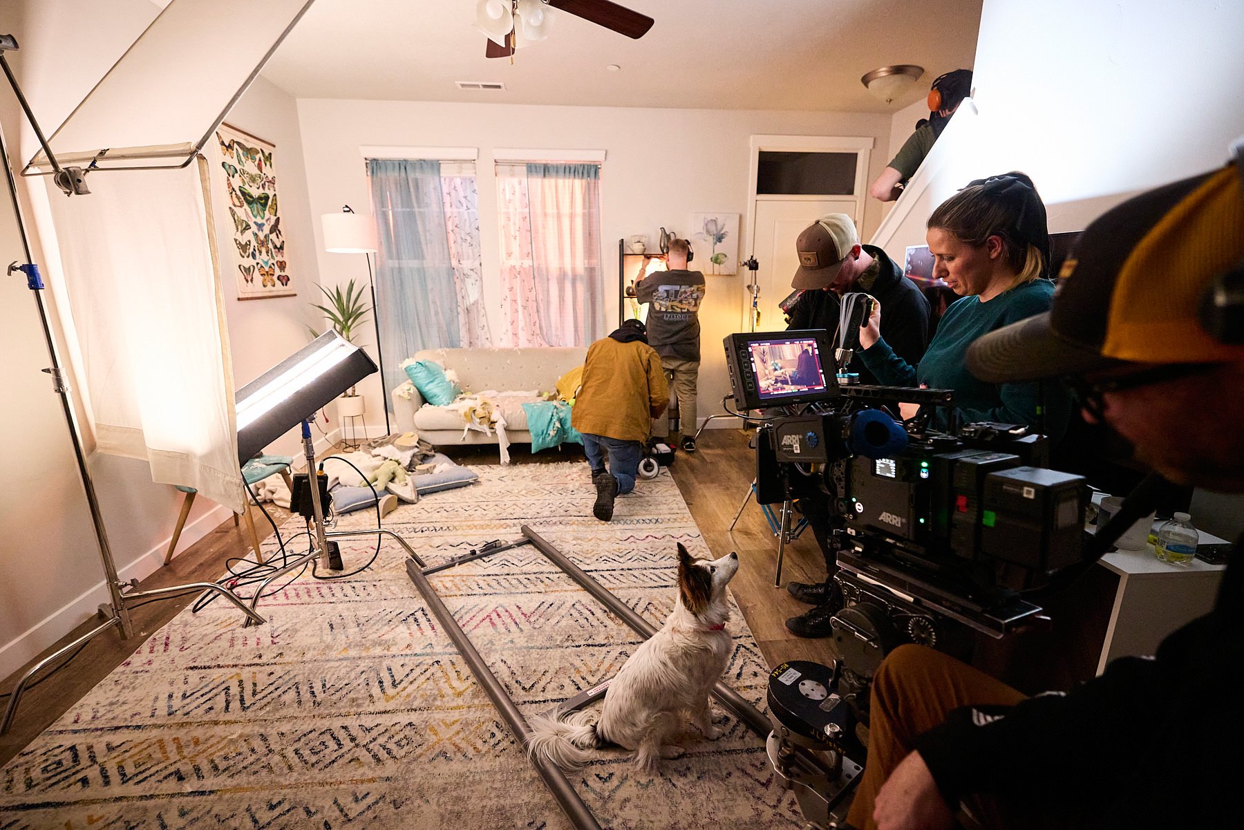 A film crew setting up a scene in a living room with a white couch, colorful pillows, and a bird design artwork on the wall. The room has a large window with curtains, and the floor is covered with a patterned rug. A camera crew is working with a pro