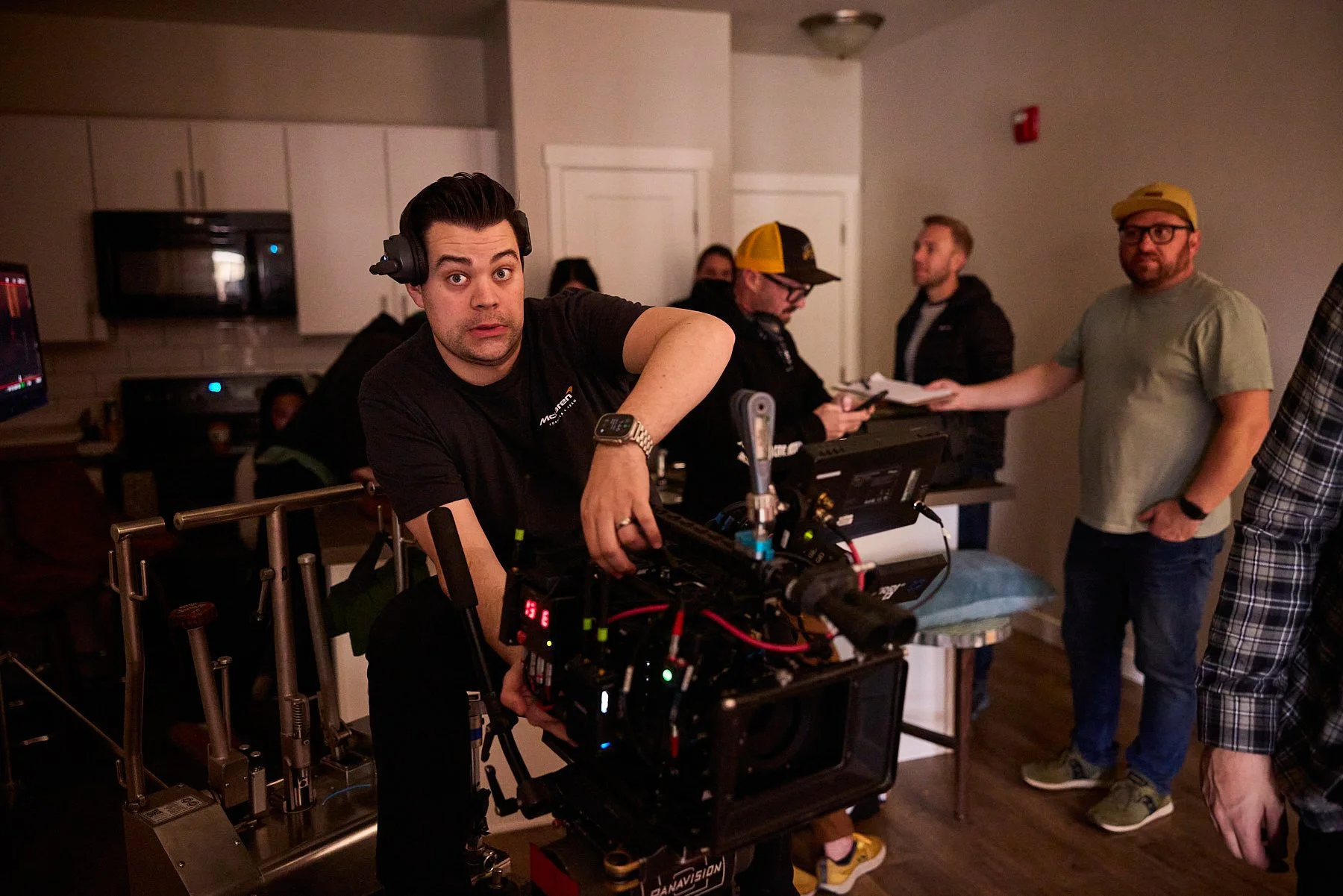 A man wearing headphones and a black T-shirt is operating a professional film camera on a dolly, with a group of people in the background on a film set.