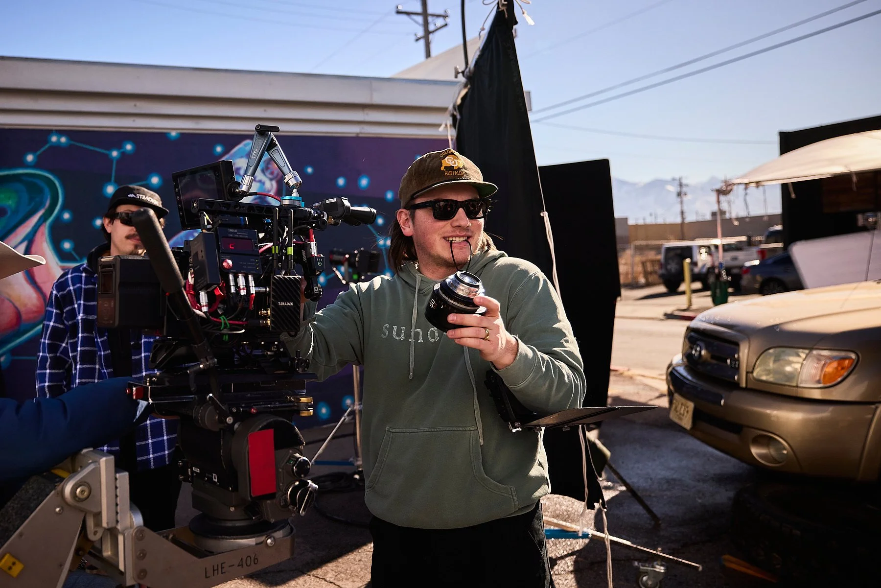 Filmmaker with camera and monitor on an outdoor film set, smiling and wearing sunglasses and a hoodie.