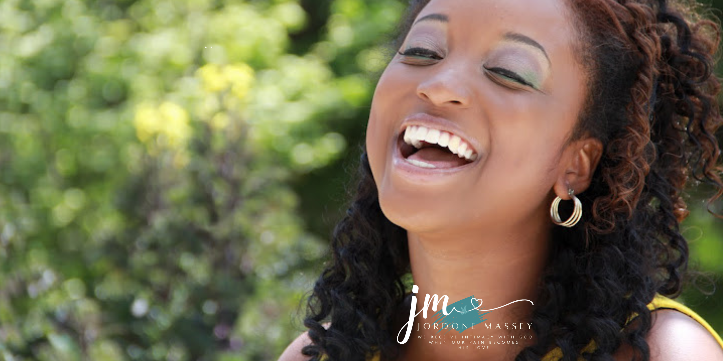 A woman with curly hair and earrings, smiling and laughing outdoors with green blurred background.