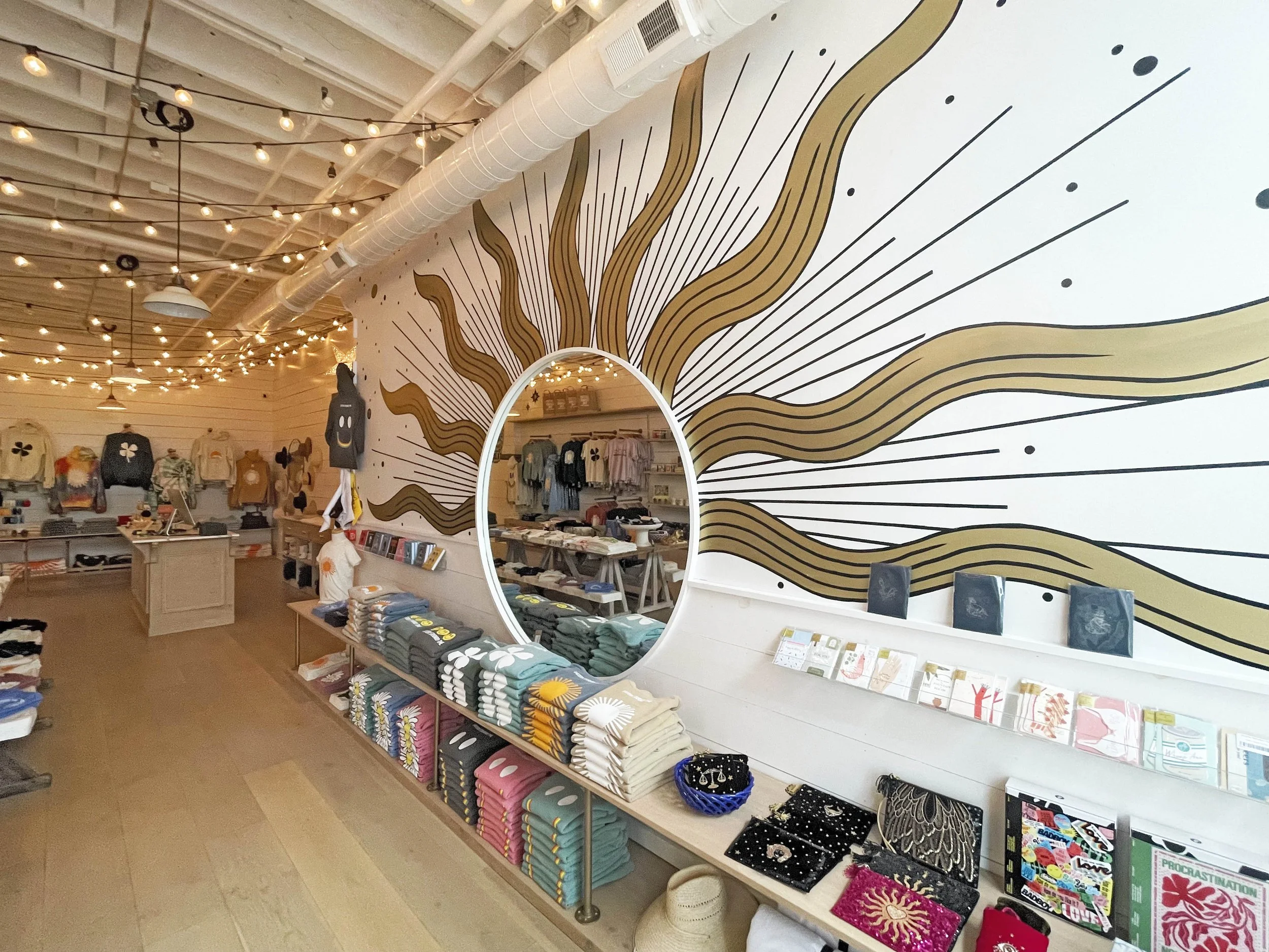 Interior of a retail store with store merchandise and a wall mural of wavy lines radiating from a mirror, decorated with string lights.