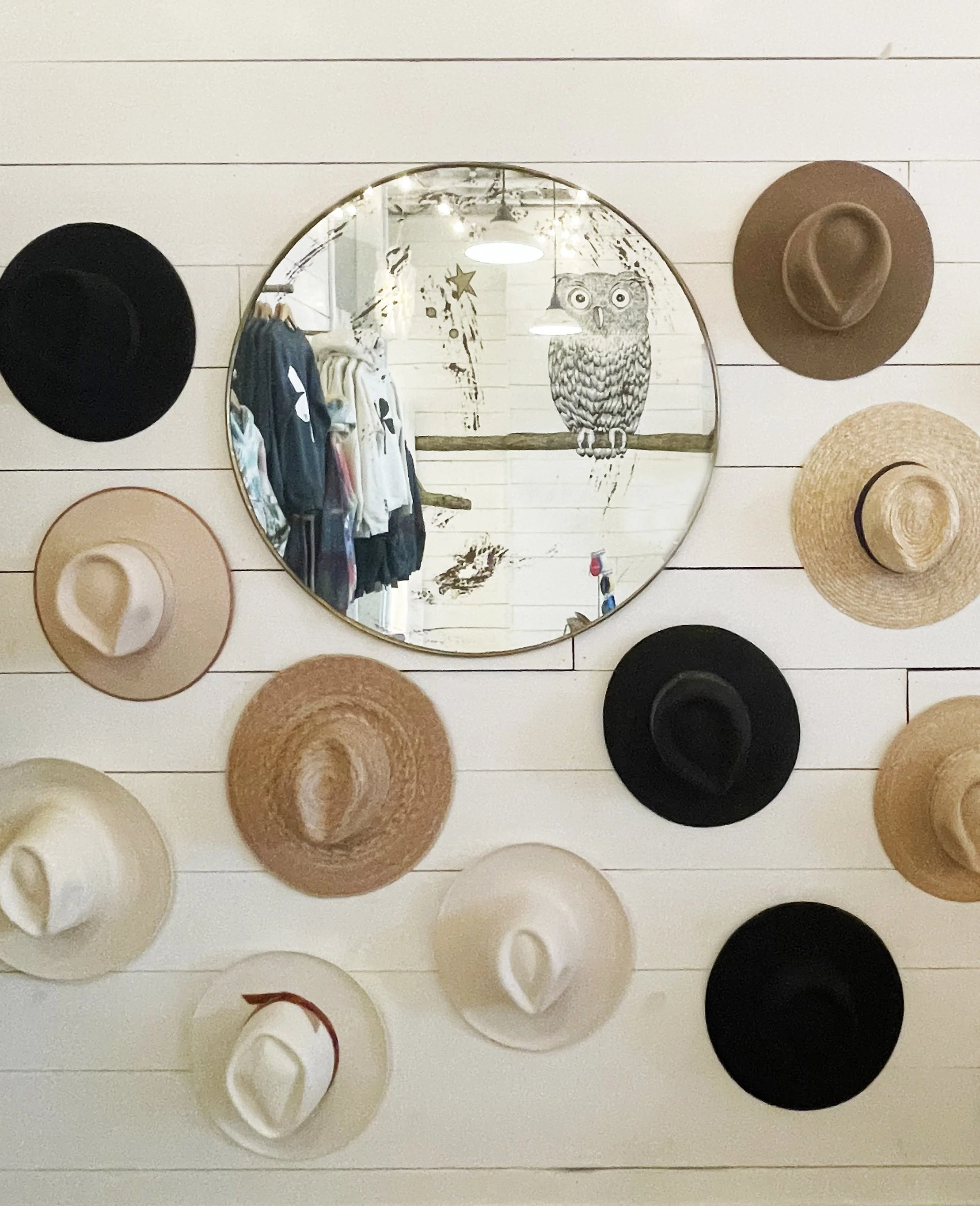 Decorative wall with a round mirror and an assortment of hats in black, beige, tan, and white colors arranged around it, with a mural of an owl and stars reflected in the mirror.