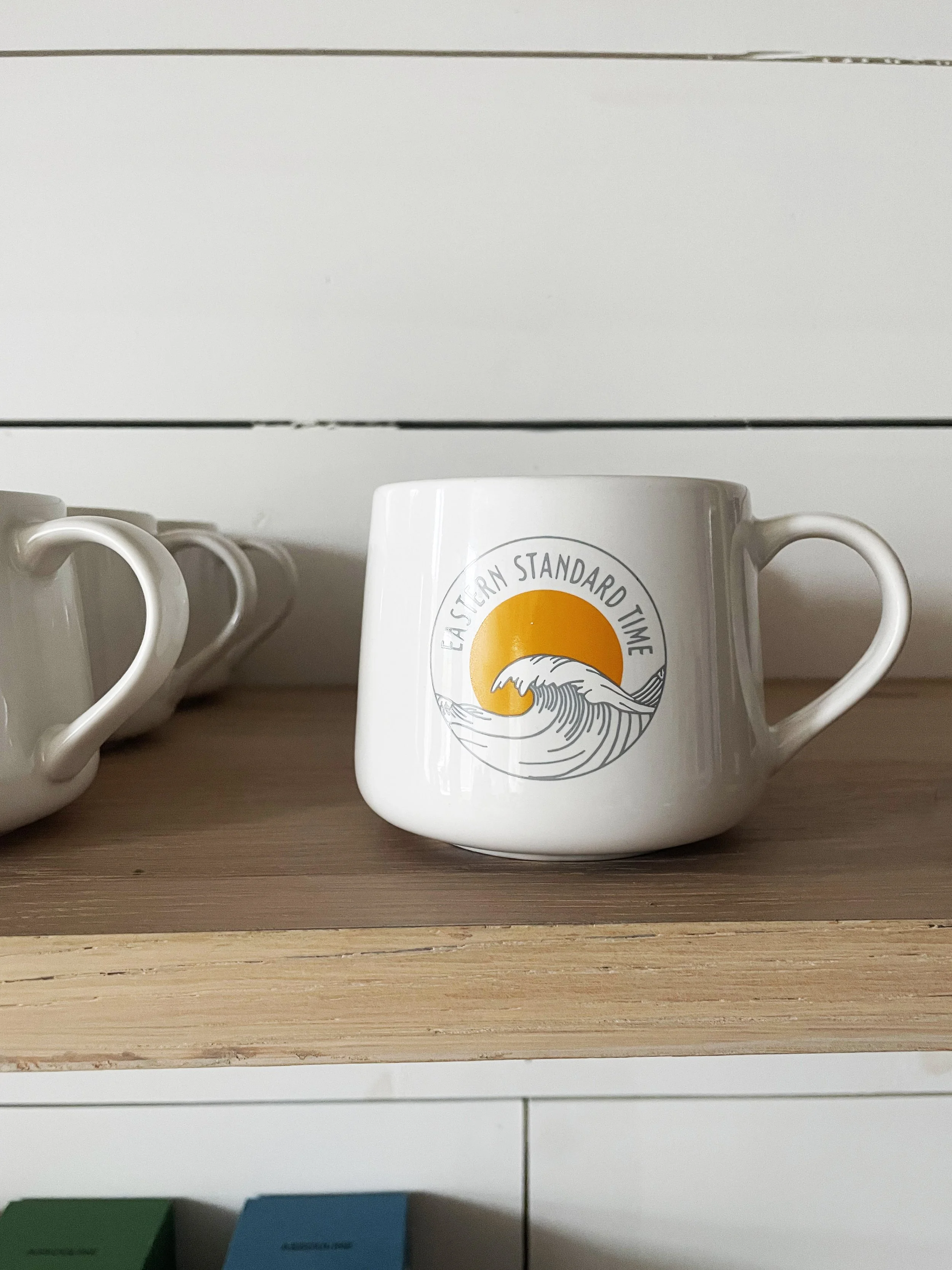 A white coffee mug with a logo that says "Eastern Standard Time" and features a sun setting behind a wave, placed on a wooden shelf, with other similar mugs to the left.