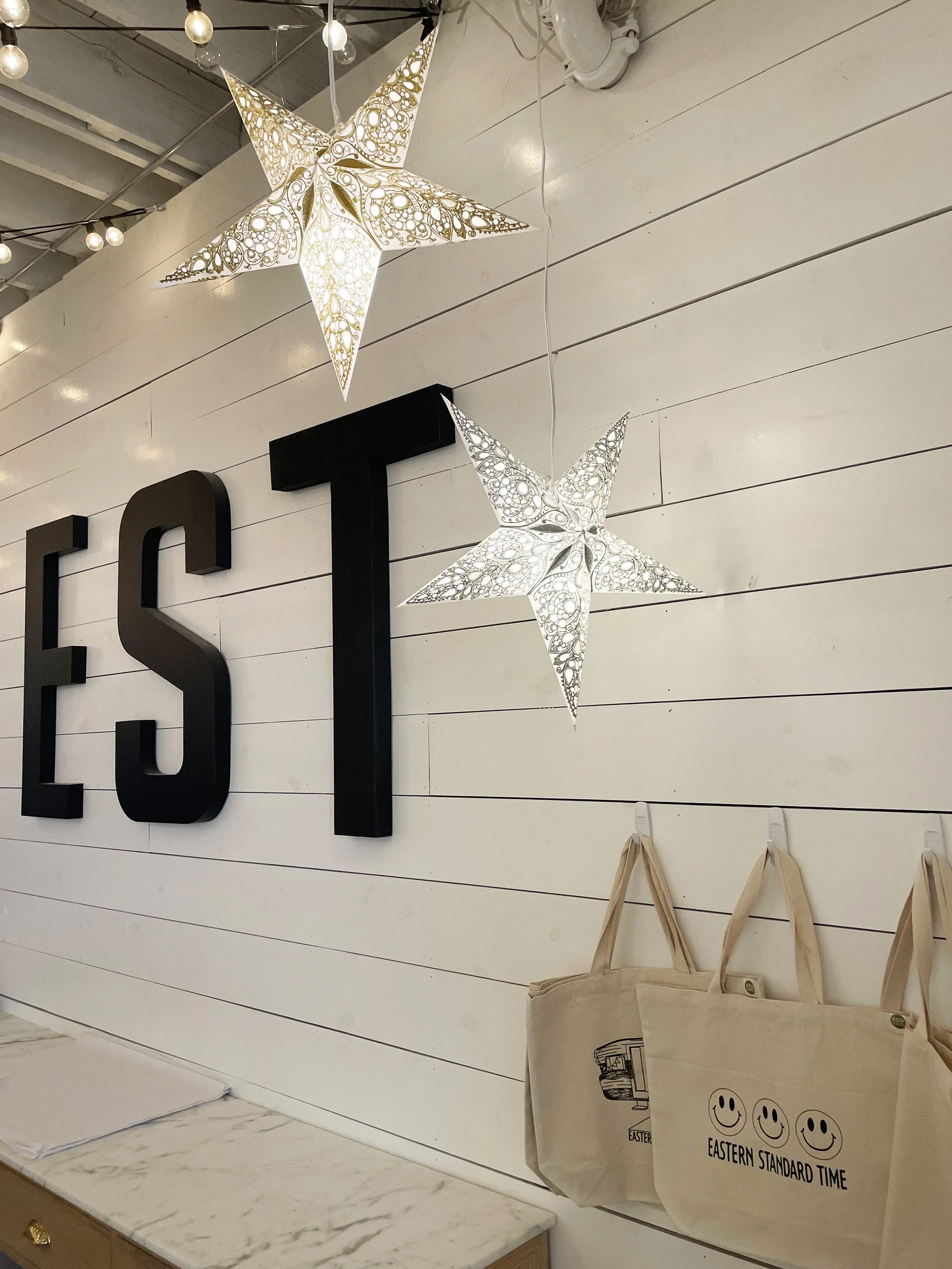 Decorative white wall with black letters spelling 'EAST'. Two illuminated star-shaped lantern decorations with intricate patterns hanging above the marble countertop. Beige tote bags with smiley faces and the text 'EASTERN STANDARD TIME' hanging on h
