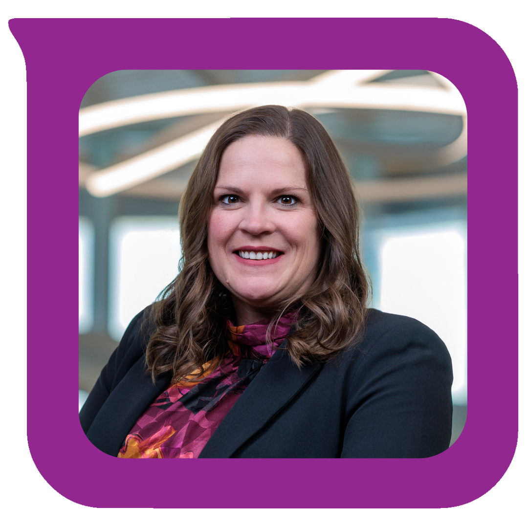 A professional woman with shoulder-length brown hair, smiling, wearing a black blazer and a purple patterned blouse, in a modern office setting.