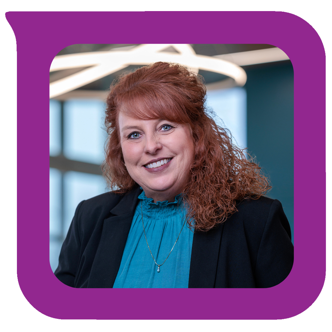 A smiling woman with red curly hair wearing a black blazer and a blue blouse, standing in a modern office with large windows and contemporary lighting.