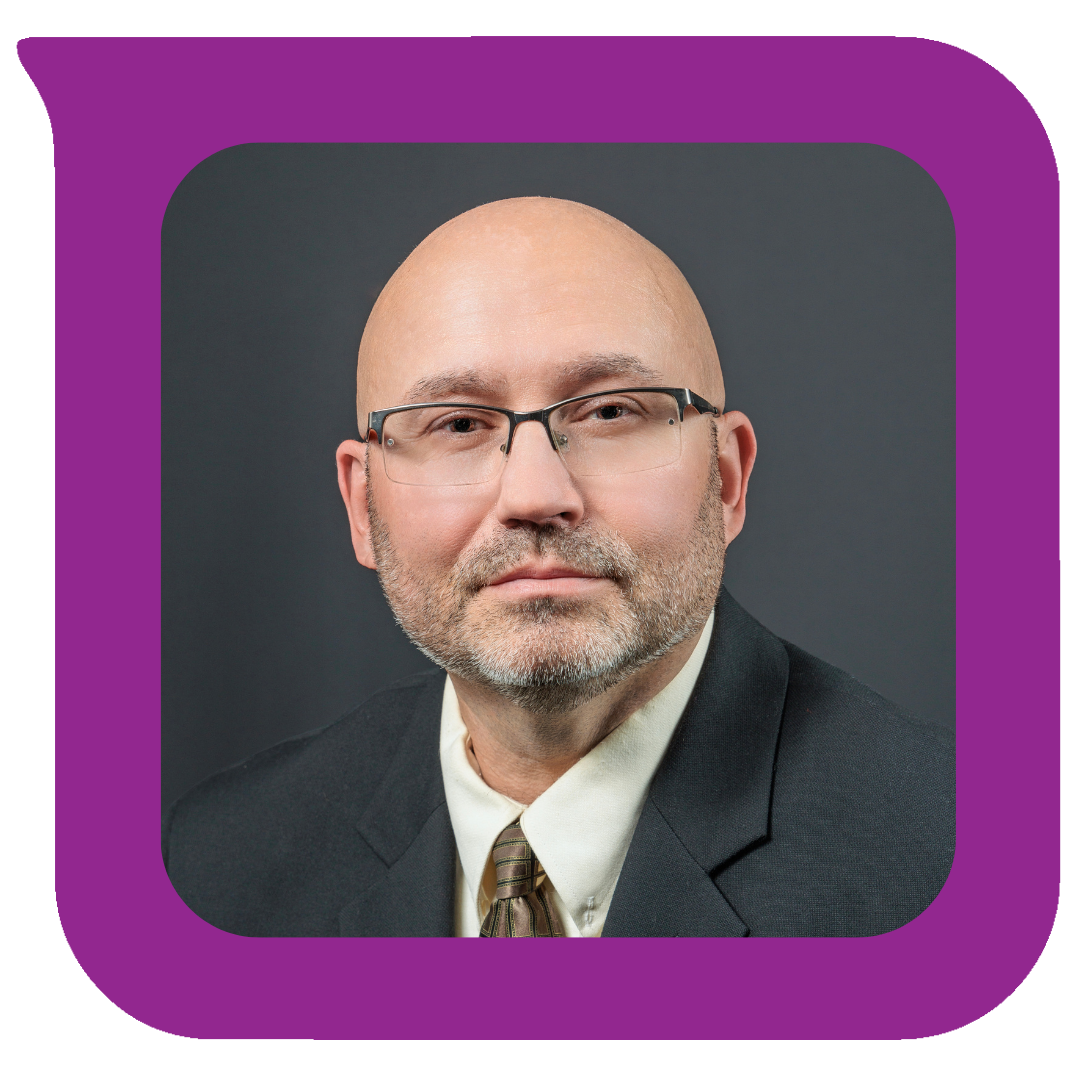 A professional headshot of a middle-aged man with a bald head, glasses, and a gray beard, wearing a black suit, white shirt, and a striped tie, against a dark gray background.