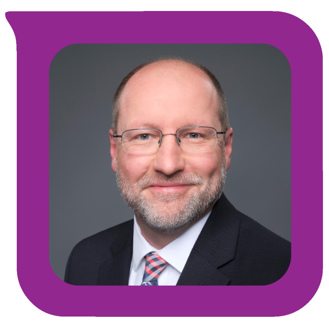 Headshot of a middle-aged man with glasses, a beard, wearing a suit and tie, smiling against a gray background.