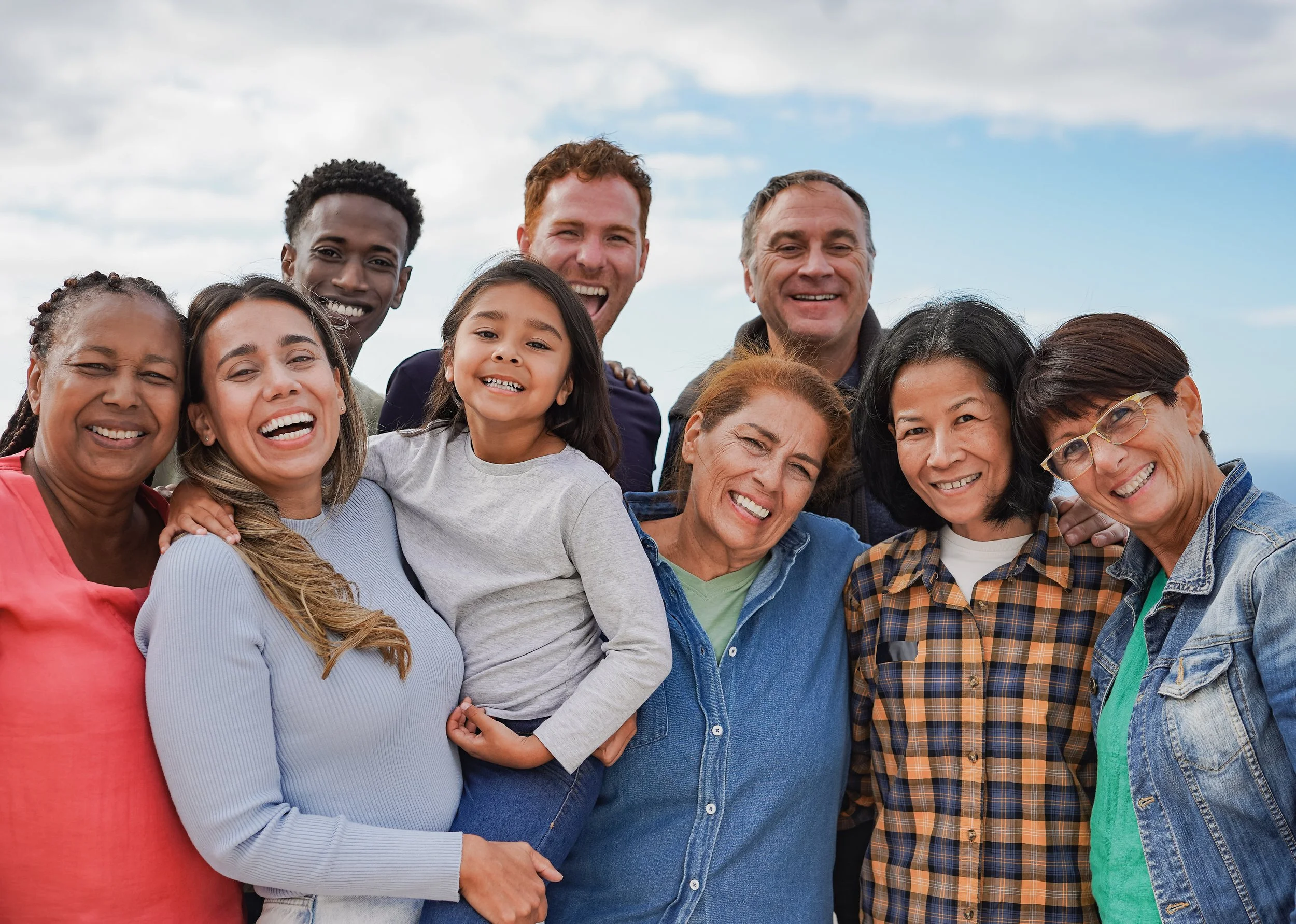A diverse group of people smiling and standing close together outdoors with a cloudy sky in the background.