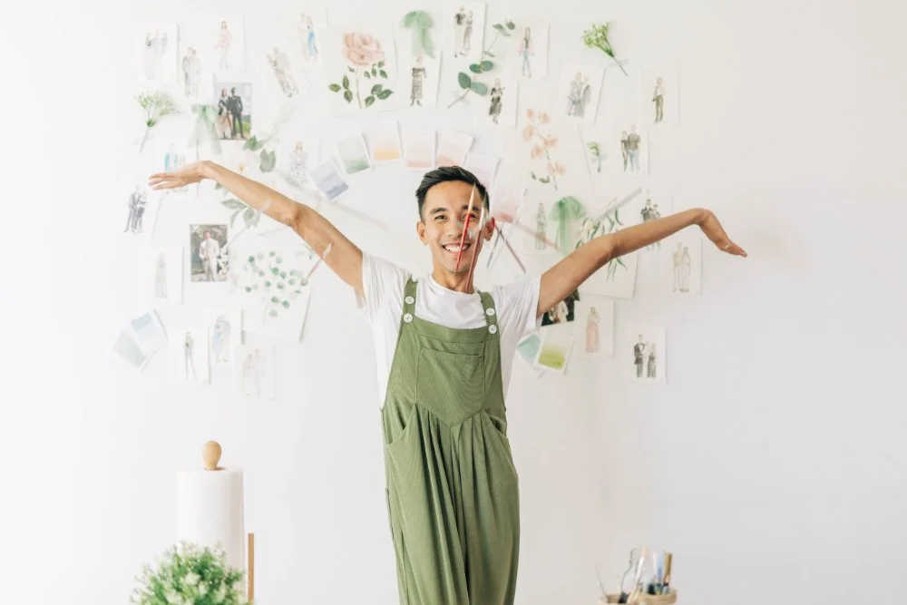Artist in green overalls poses playfully in front of a wall of sketches, swatches, and florals—bright, creative studio energy that fits couples planning an eco-friendly wedding with intentional, experience-first details.