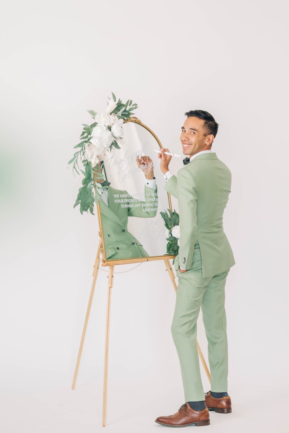 Artist in a sage suit hand-letters a “Unplugged Ceremony” mirror sign on an easel, framed with white blooms and greenery—elegant ceremony signage inspiration for an eco-friendly wedding with a minimal, modern vibe.