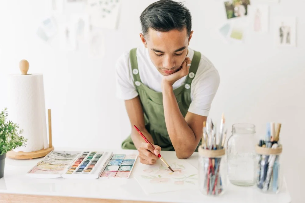 Artist in green overalls and a white tee paints at a tidy workstation with jars of brushes, watercolor pans, and paper laid out. Soft natural light and minimal styling show a calm, elevated process for creating quick guest portraits.
