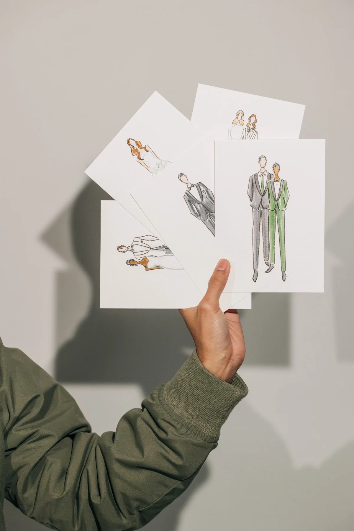 Hand holding a fan of watercolor fashion portrait cards—stylized couples and outfits—against a soft gray wall with dramatic shadow.