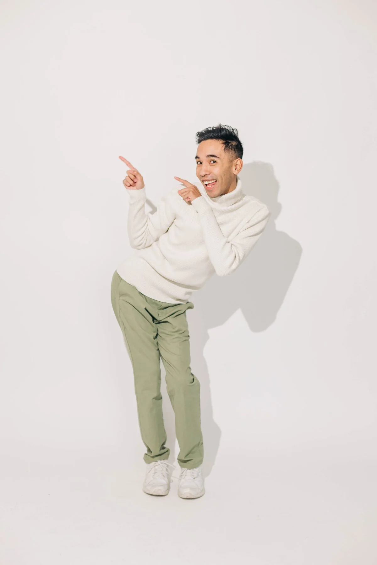Playful studio portrait of a smiling man in a cream sweater and sage pants pointing off-frame on a clean white backdrop.