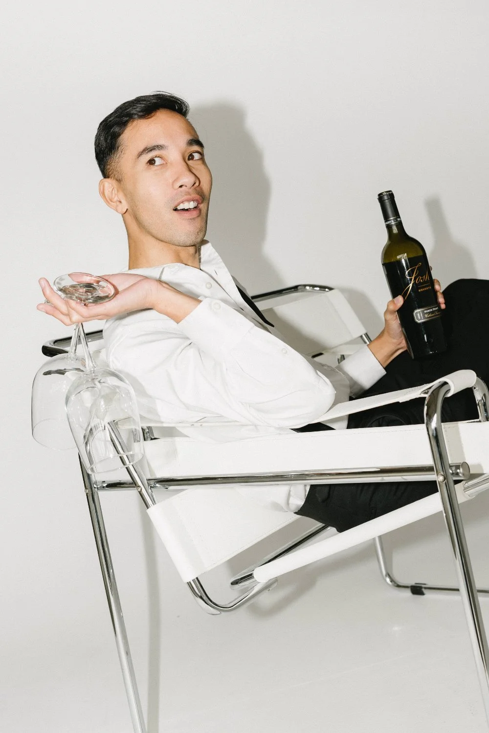 A stylish person lounges in a white chair, balancing an inverted wine glass while holding a wine bottle—playful, modern imagery for wine country weddings and winecolor guest portraits.