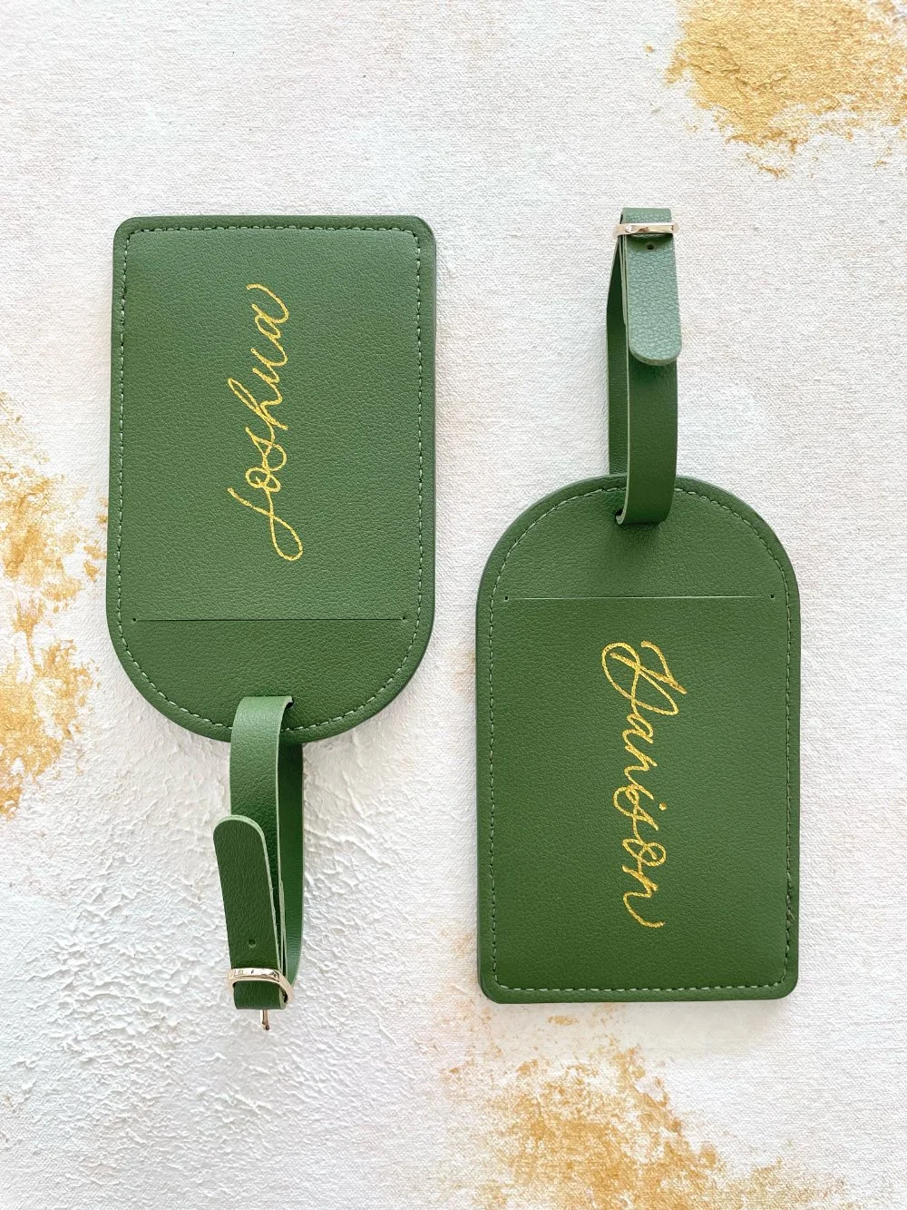  Two forest-green leather luggage tags with gold handwritten names on a textured background—an elevated favor idea for  live calligraphy and on-the-spot engraving  at weddings and events. 