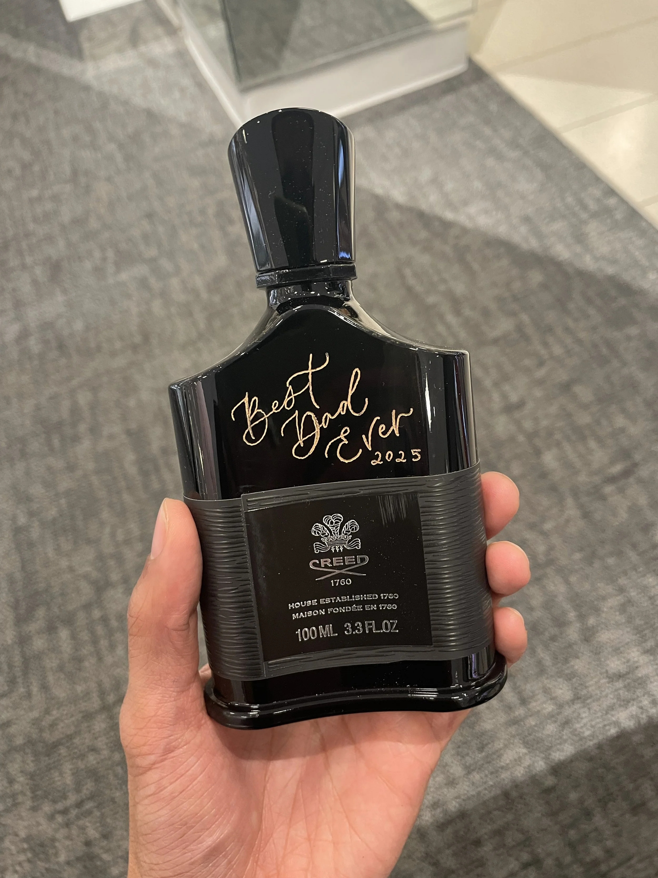  Hand holds a black Creed bottle with gold handwritten script reading “Best Dad Ever 2025”—a premium personalized gift created through  live calligraphy and on-the-spot engraving . 
