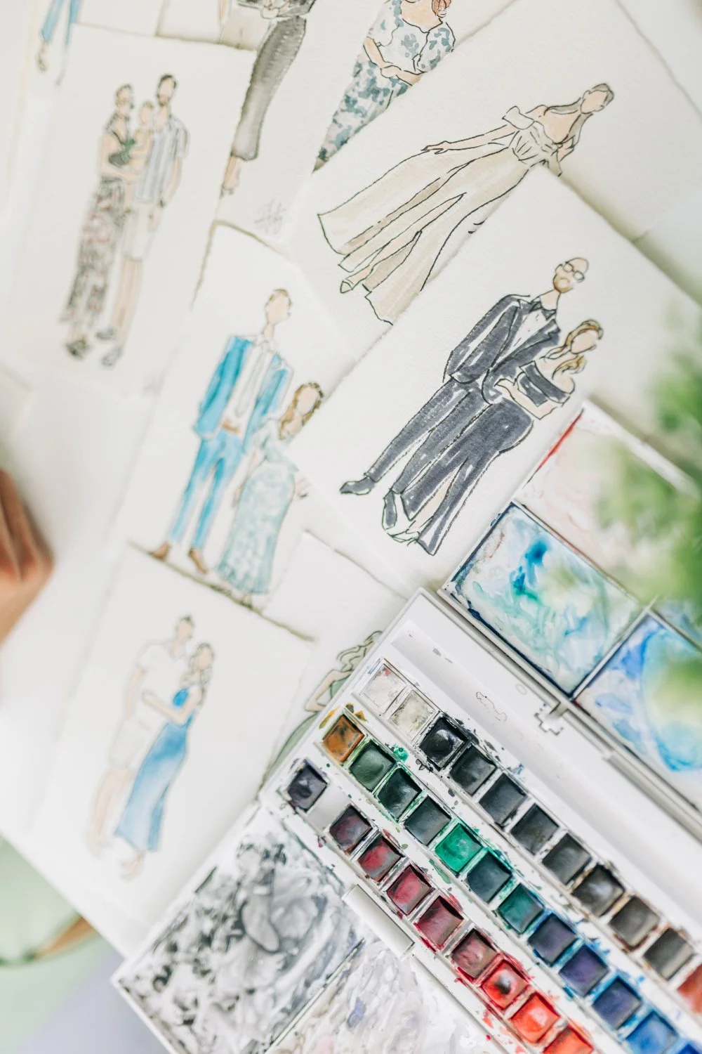 Flat lay of finished watercolor fashion portraits on textured paper beside a well-loved paint palette—an elevated keepsake idea for an eco-friendly wedding that replaces disposable favors with art guests actually want to keep.