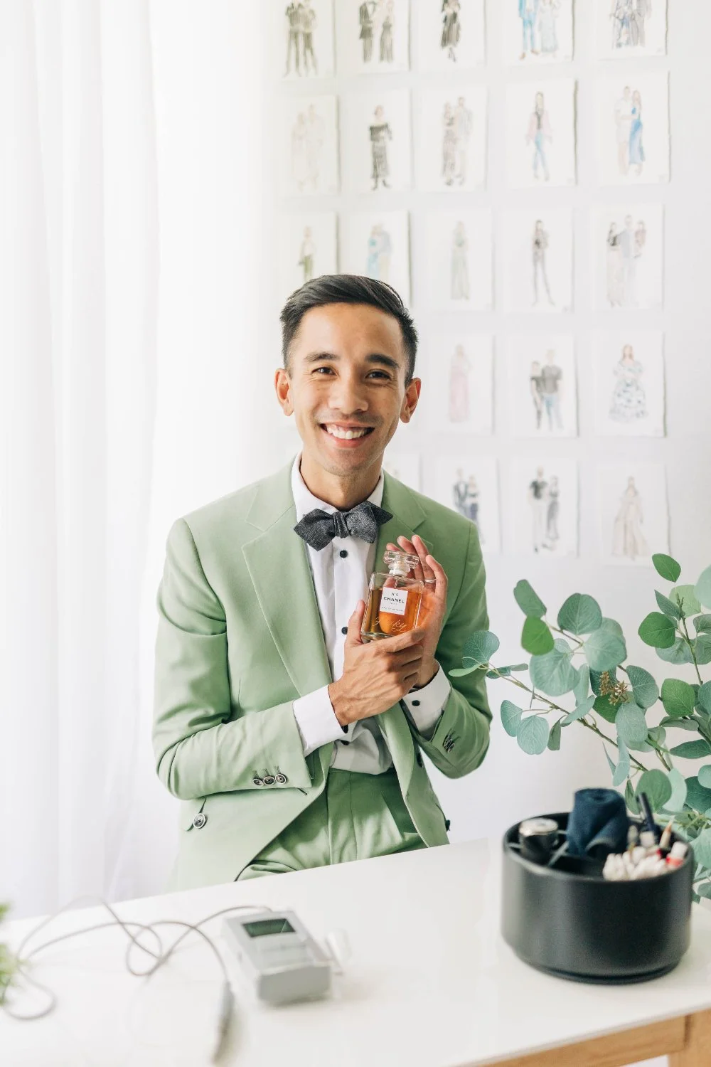 Artist in a sage suit smiles while holding a perfume bottle at a clean workstation, with greenery and a wall of finished guest portraits behind him.